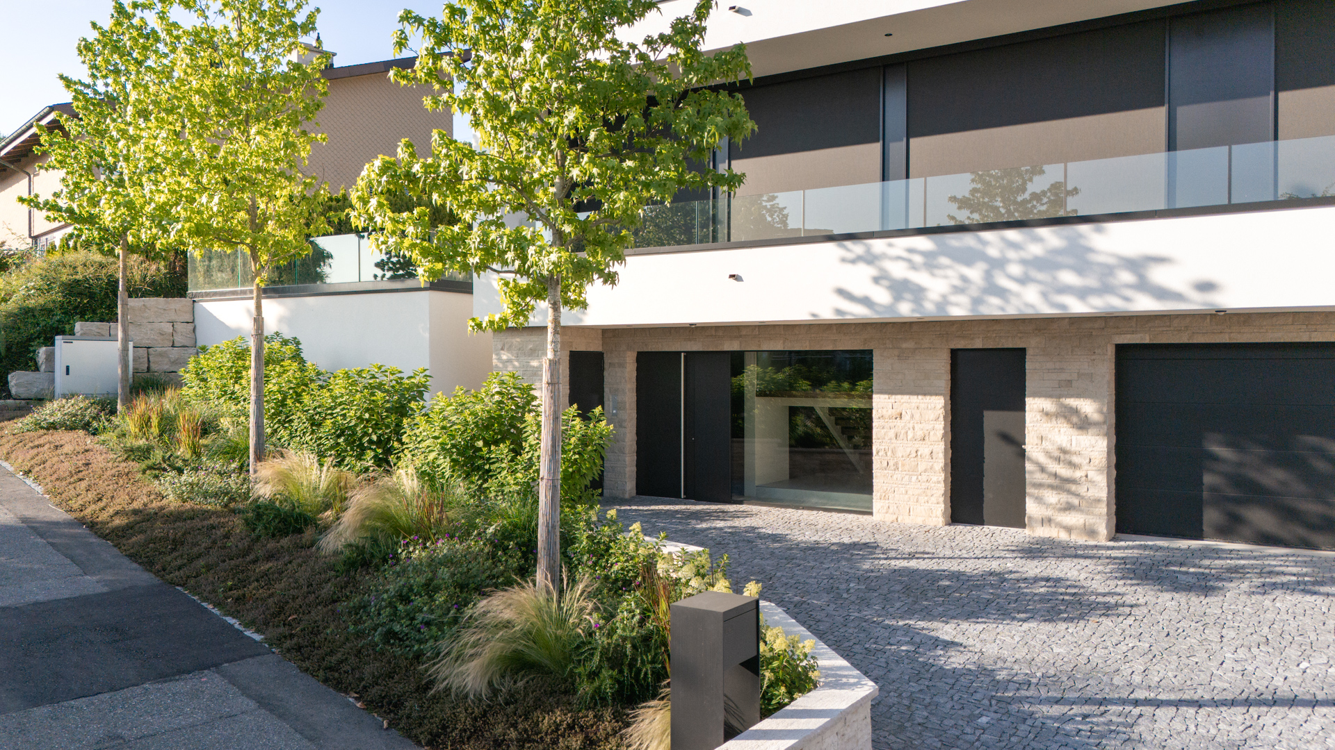 Auffahrt zu einem modernen Luxus Haus mit Stauden, Sträuchern und hochstämmigen Bäumen. 