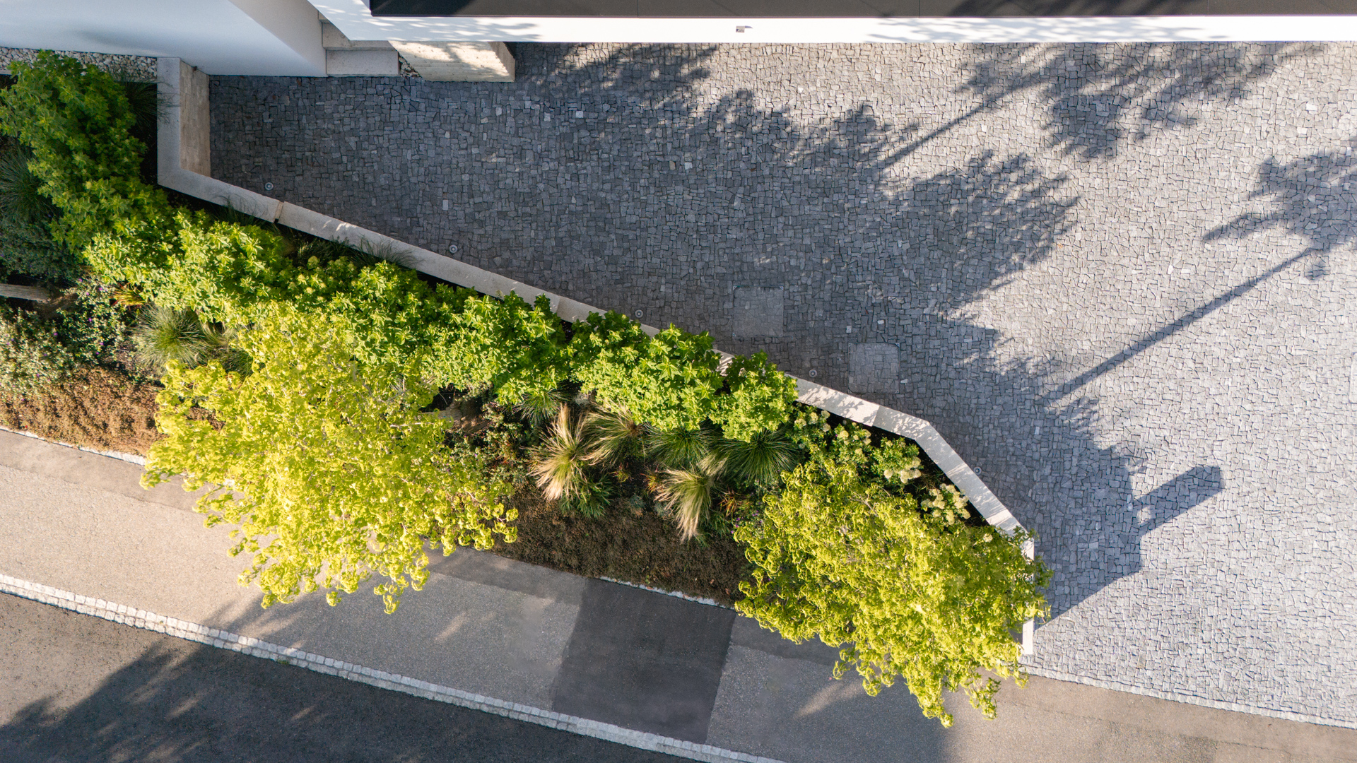 Drohnenaufnahme der Einfahrt eines Privathauses mit schöner Bepflästerung und Blumenbeet mit hochstämmigen Bäumen welche Schatten werfen. 