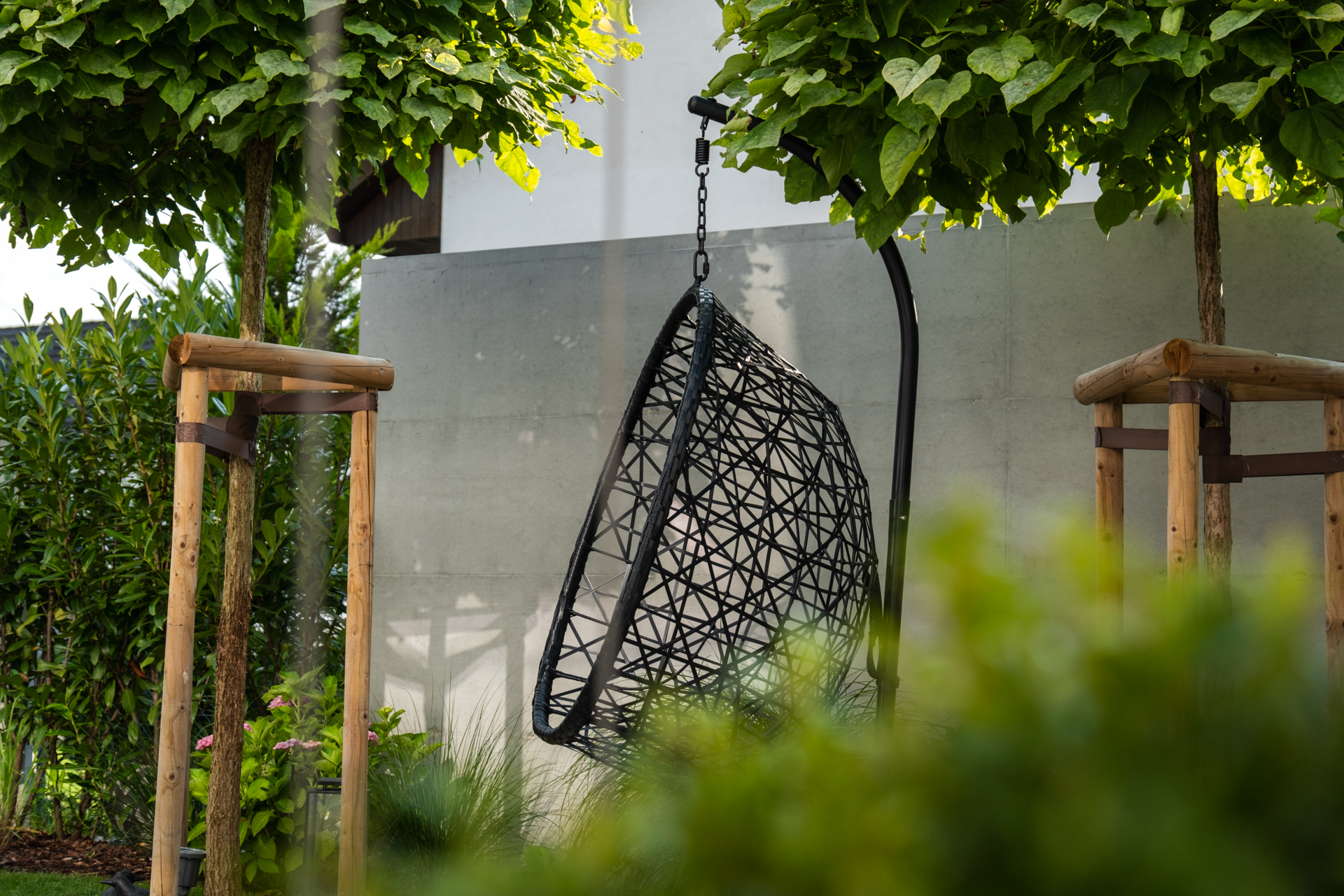 Moderner Korbhängesessel unter hochstämmigen Bäumen im Schatten. 