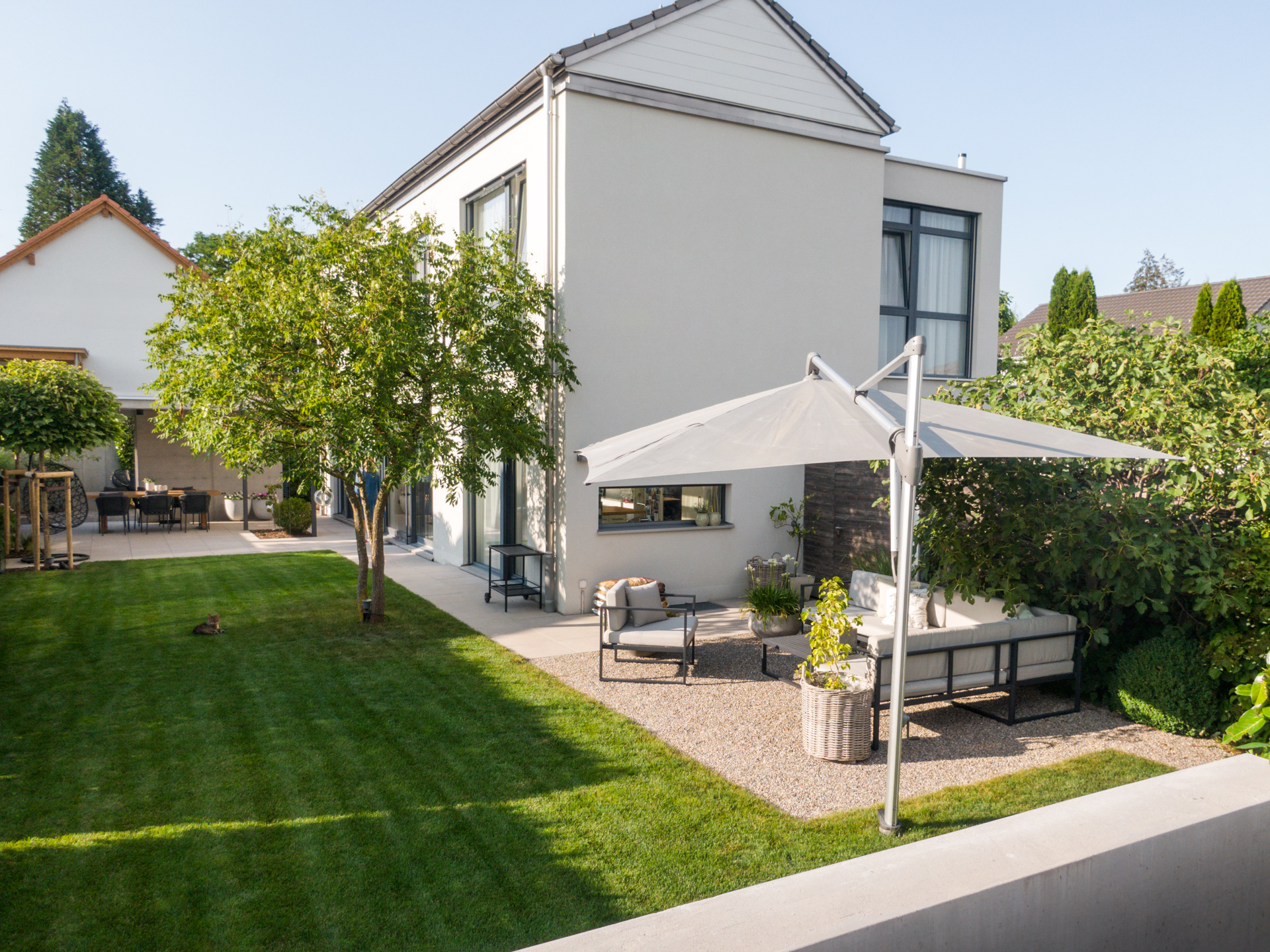 Modernes Wohnhaus mit Kiesplatz im Garten, grossen Rasenflächen und einer Gartenlounge unter einem Sonnenschirm. 