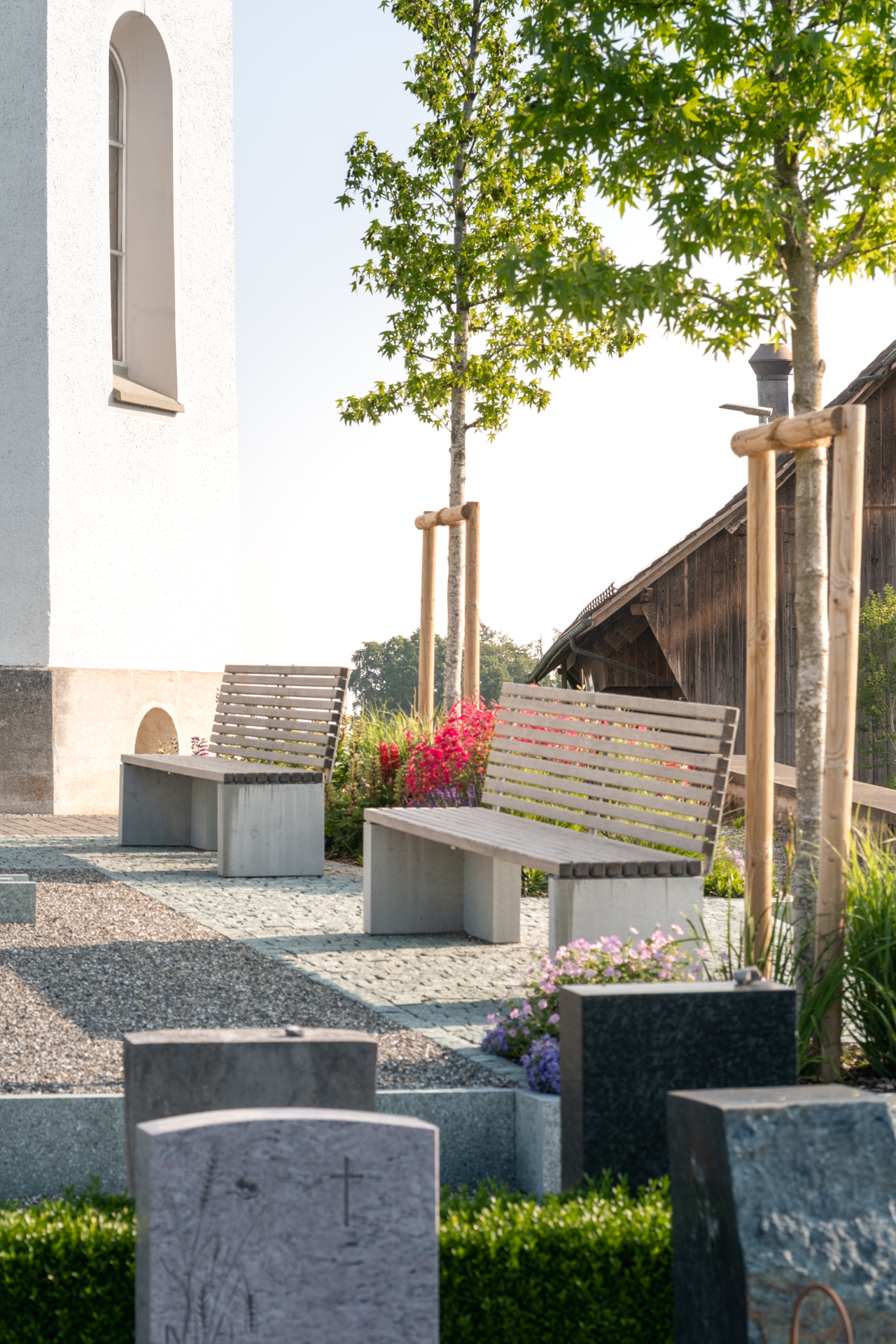 Holzsitzbänke auf einem Friedhof umgeben von Blumenbeeten und hochstämmigen Bäumen. 