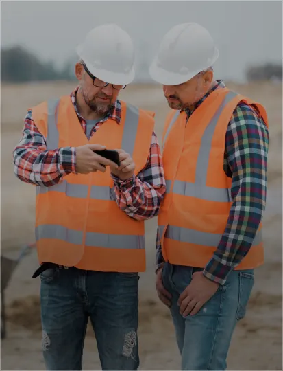 Two men wearing construction vests and helmets, one demonstrating something on a mobile phone.