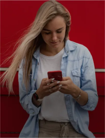 A woman stands in front of a red wall, looking at her phone, absorbed in her device.