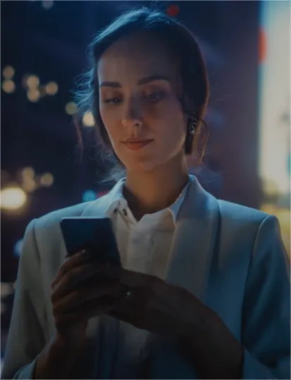 A woman in a business suit checks her phone at night, illuminated by the screen's glow.