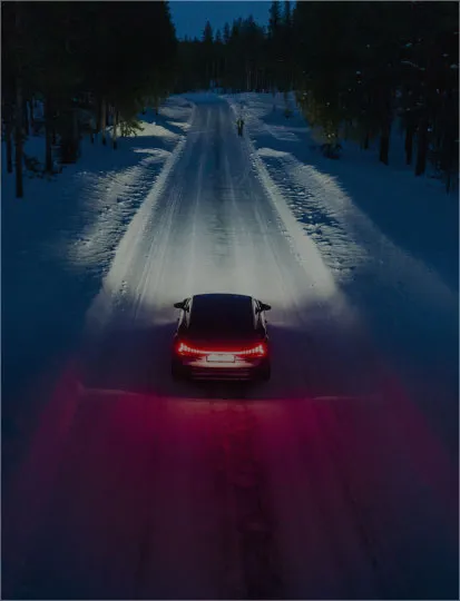 A car navigates a snowy road at night, illuminated by headlights against the dark, wintry landscape.