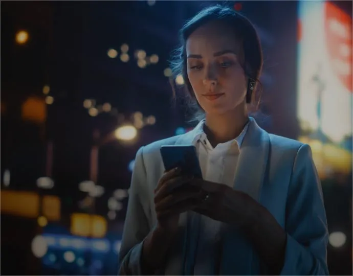 A woman in a business suit checks her phone at night, illuminated by the screen's glow.