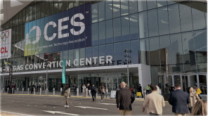 The exterior of the Las Vegas Convention Center during CES (Consumer Electronics Show), featuring a large banner for the event. Attendees walk toward the glass-fronted entrance where "Vegas Convention Center" is visible.