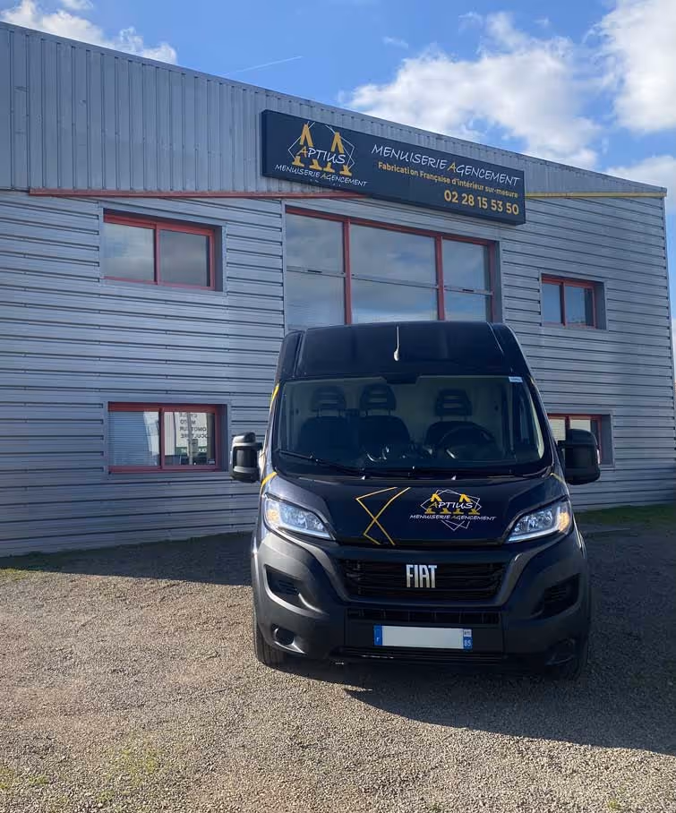 Fourgon Fiat noir garé devant un bâtiment gris avec une enseigne Aptius Menuiserie Agencement affichant un numéro de téléphone.