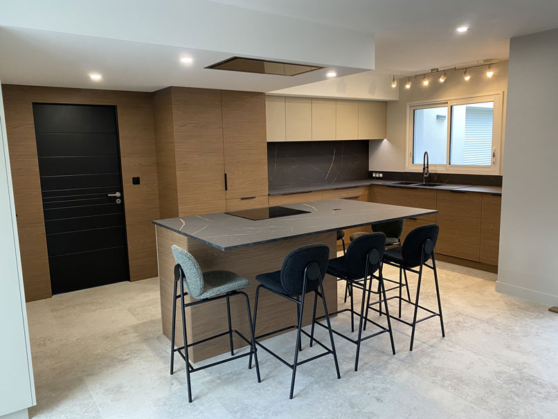 Vue d’ensemble de la cuisine aménagée avec îlot central, façades bois, meubles beige et plan de travail foncé veinage cuivré, éclairage encastré moderne.