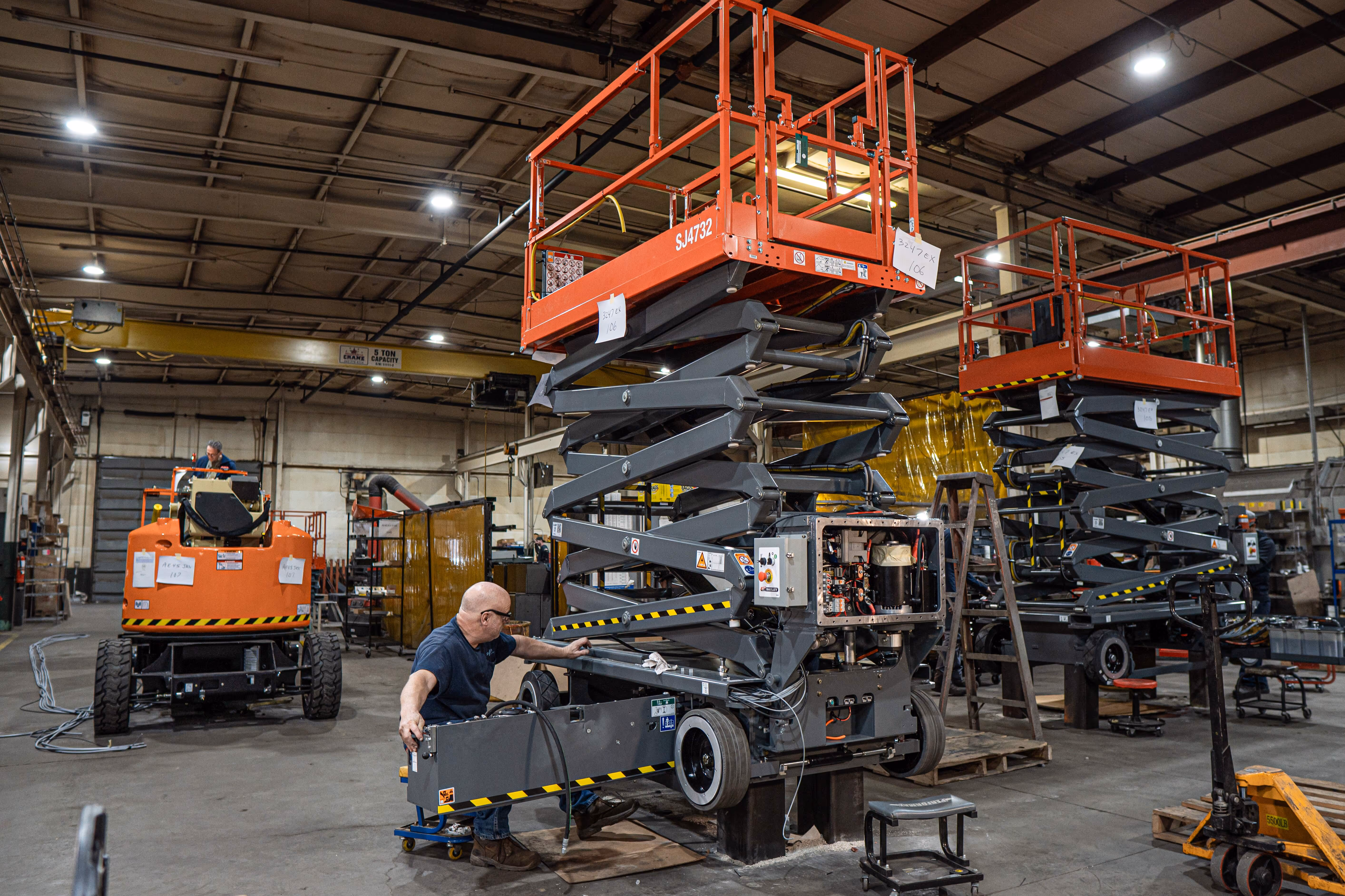 Worker seated on a rolling stool repairing the base machinery of a lifted red scissor lift in an industrial warehouse.