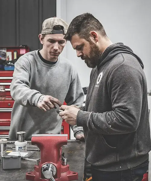 Two men working together on a small object in a workshop with tools and a red vise on the bench.