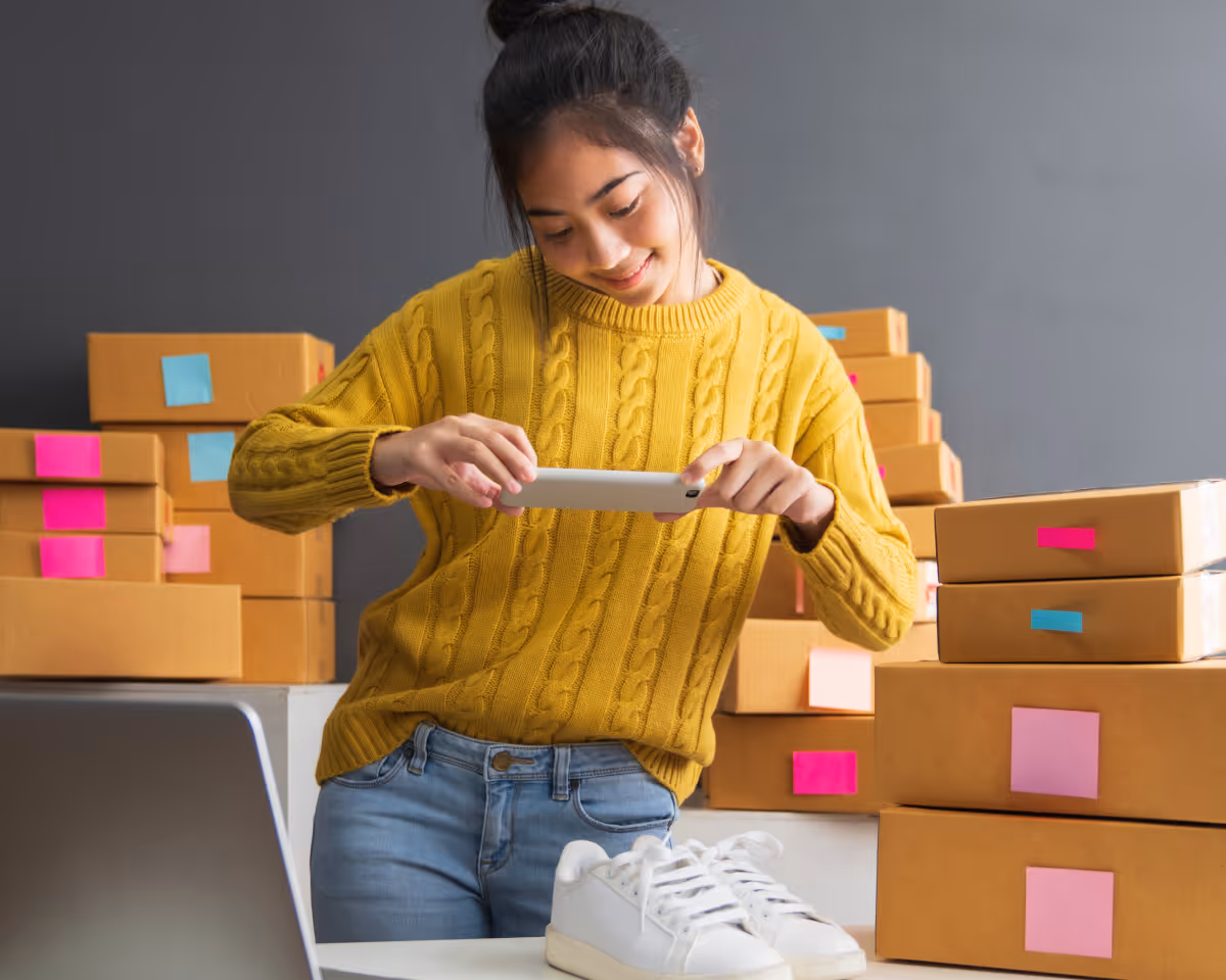 Young woman in a yellow sweater taking a photo of white sneakers with a smartphone, surrounded by cardboard shipping boxes with colorful labels.