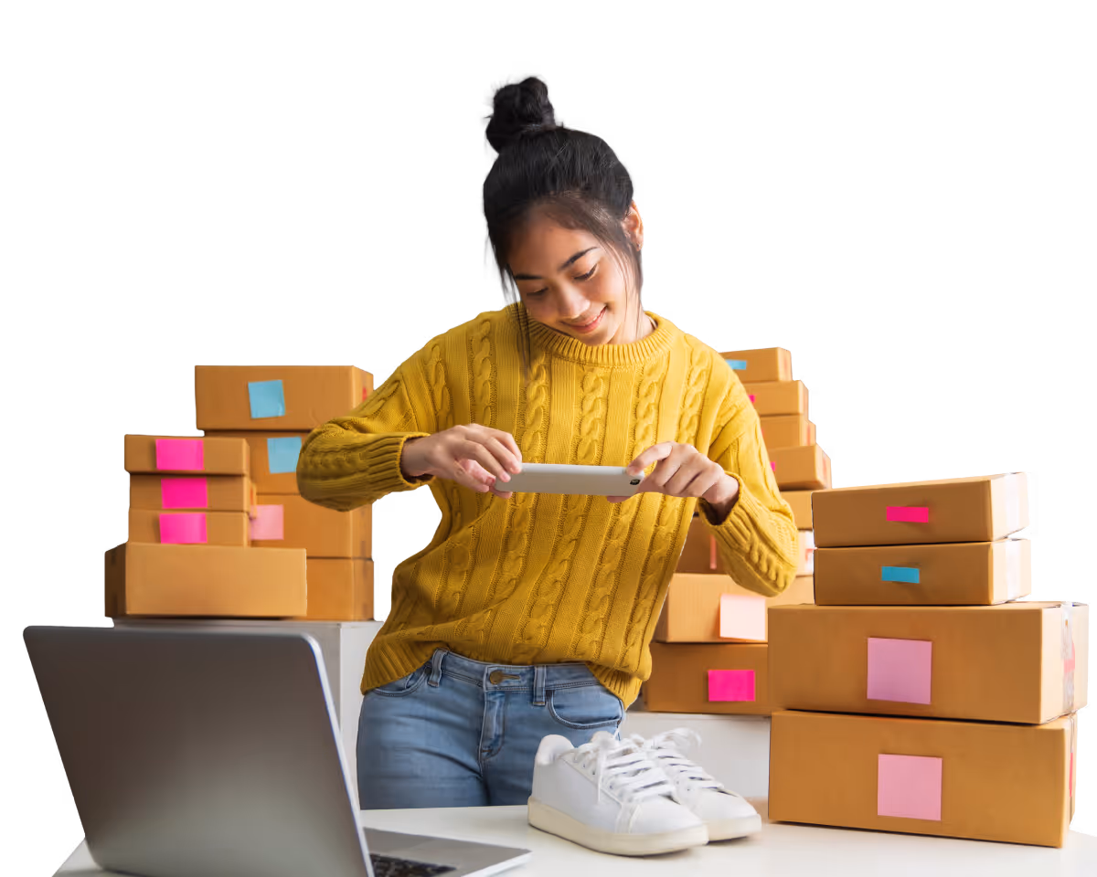 Woman in yellow sweater taking a photo of white sneakers with a smartphone surrounded by cardboard shipping boxes with colorful labels.