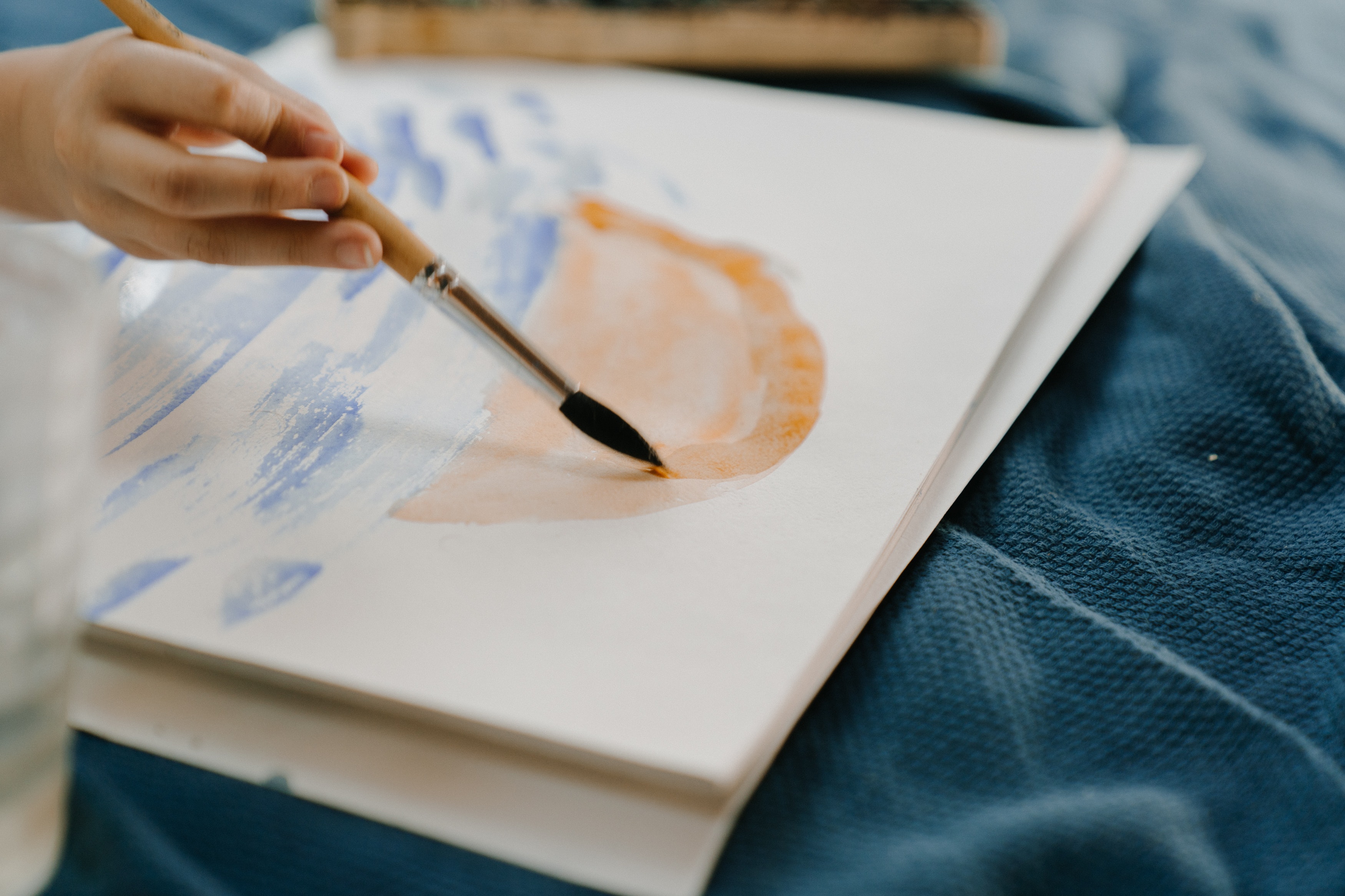 A close-up of a hand holding a paintbrush, making loose abstract shapes in blue and orange on a canvas resting on soft blue fabric
