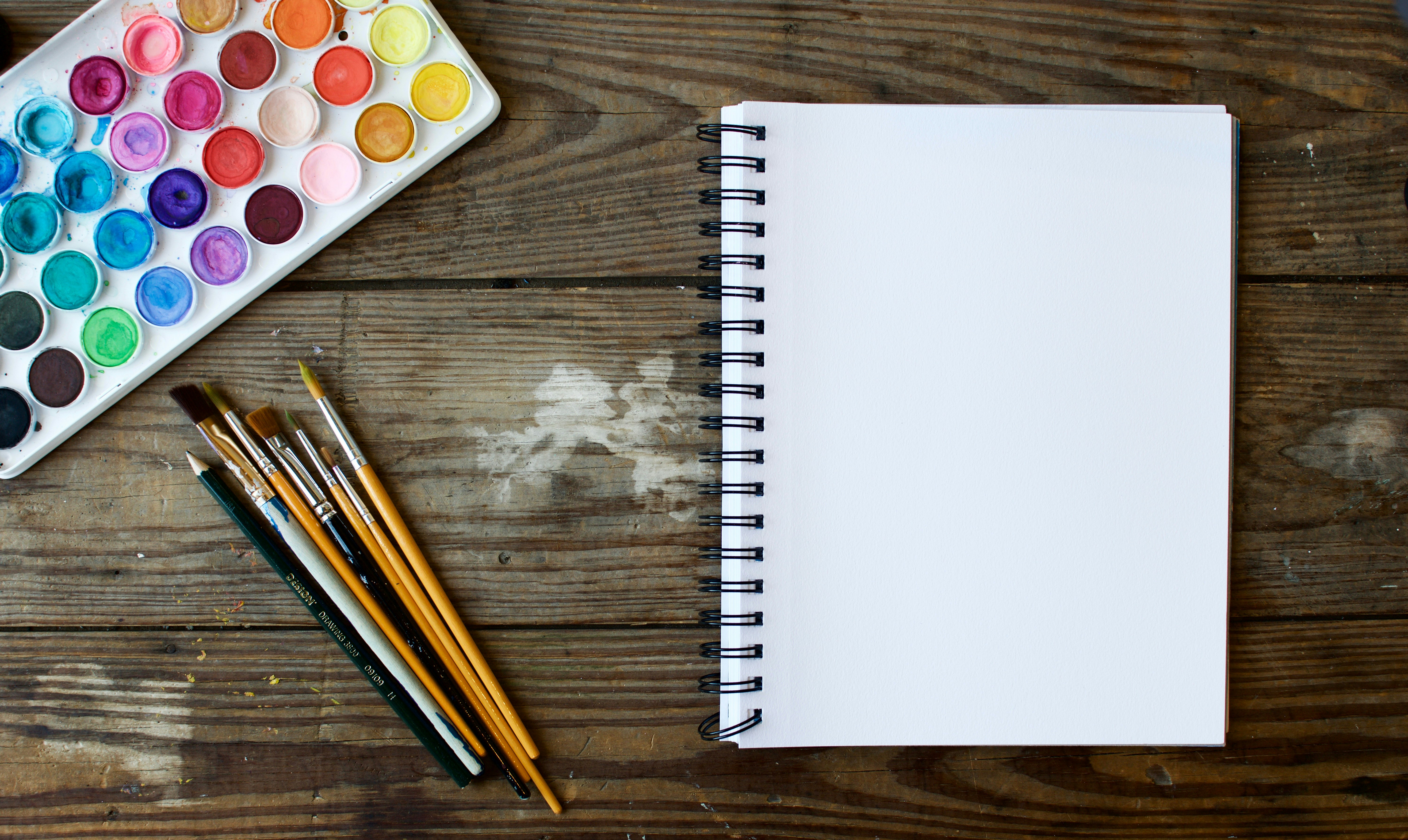 A blank open sketchbook beside a watercolour palette and several paintbrushes on a warm wooden table, quiet and inviting
