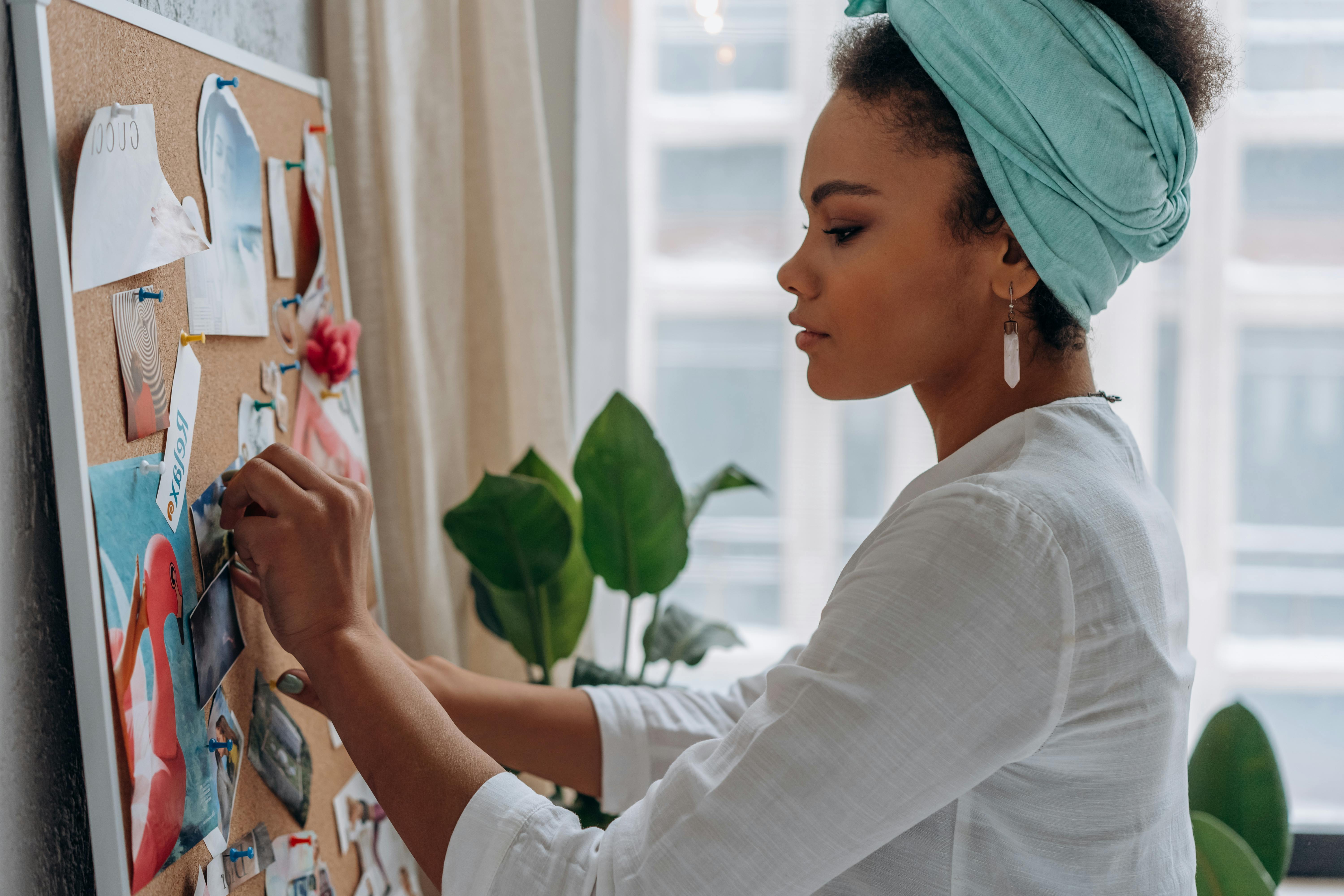 A woman in a white top standing in profile, carefully pinning an image to a cork board covered with photographs and papers, a large green plant visible beside her