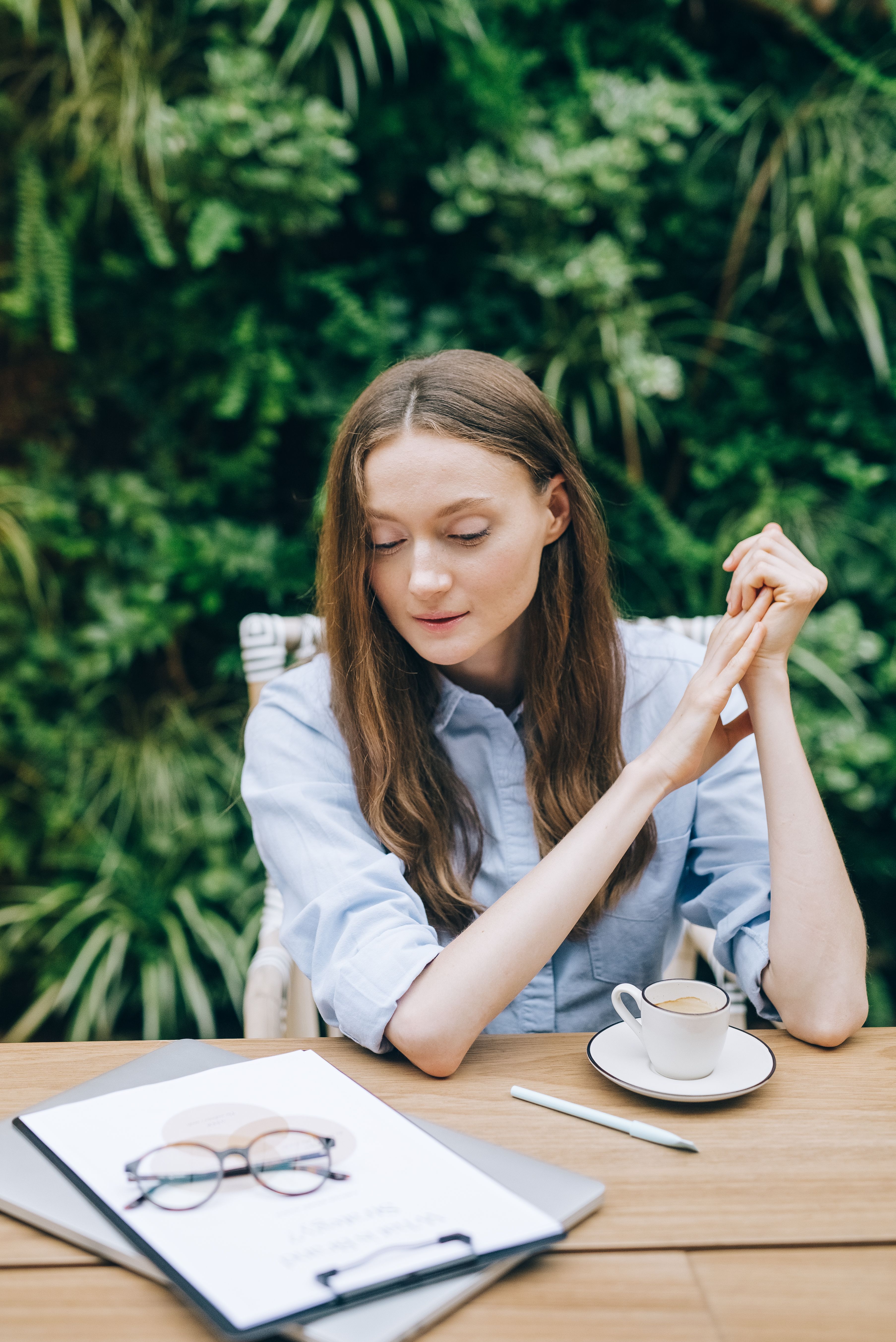 A woman in a light blue shirt sitting at an outdoor table with her hands pressed together in front of her, gaze turned downward, lush green foliage behind her