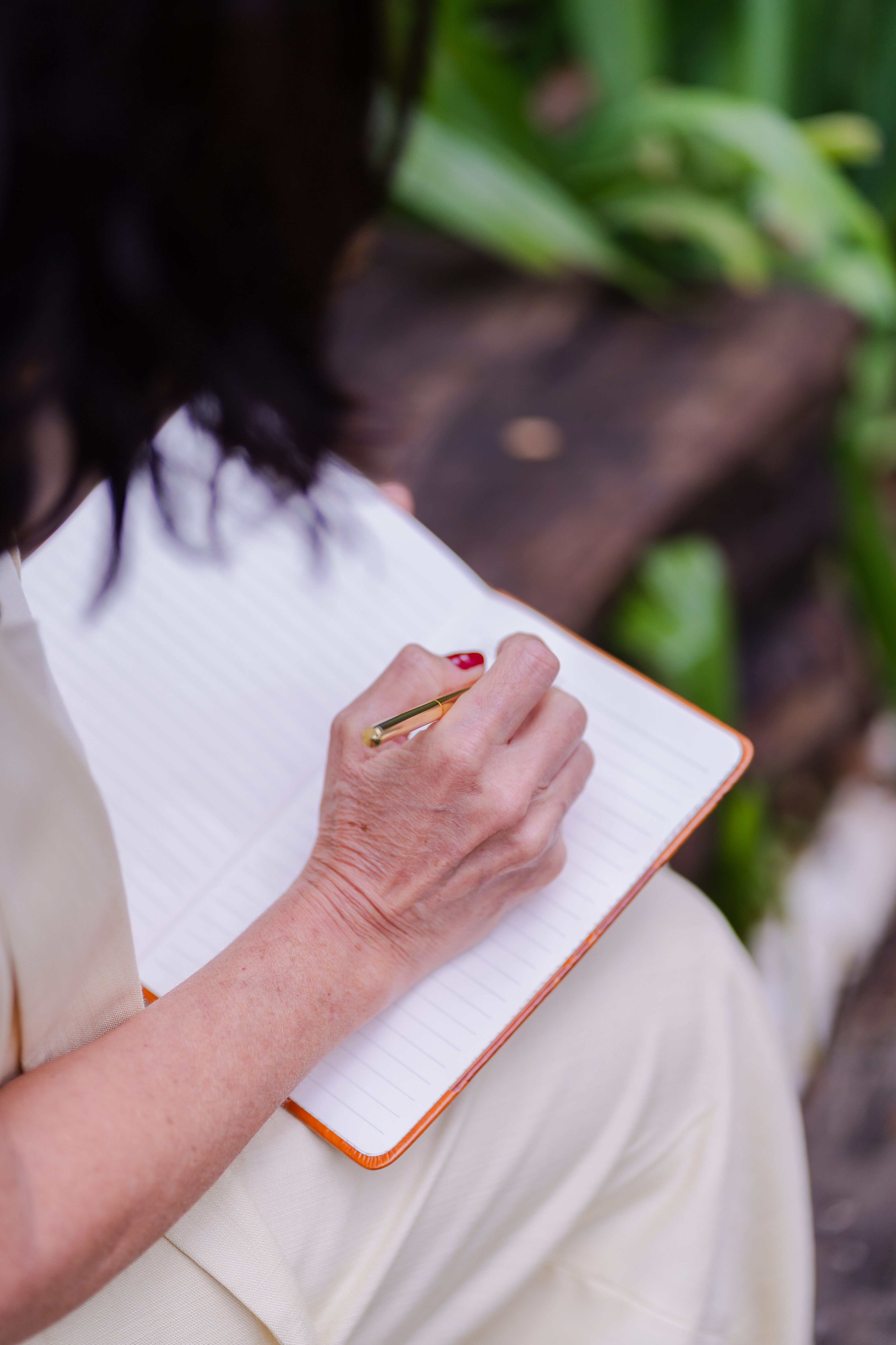 A close-up of a woman's hand holding a gold pen poised over a blank lined notebook, soft green plants visible in the background