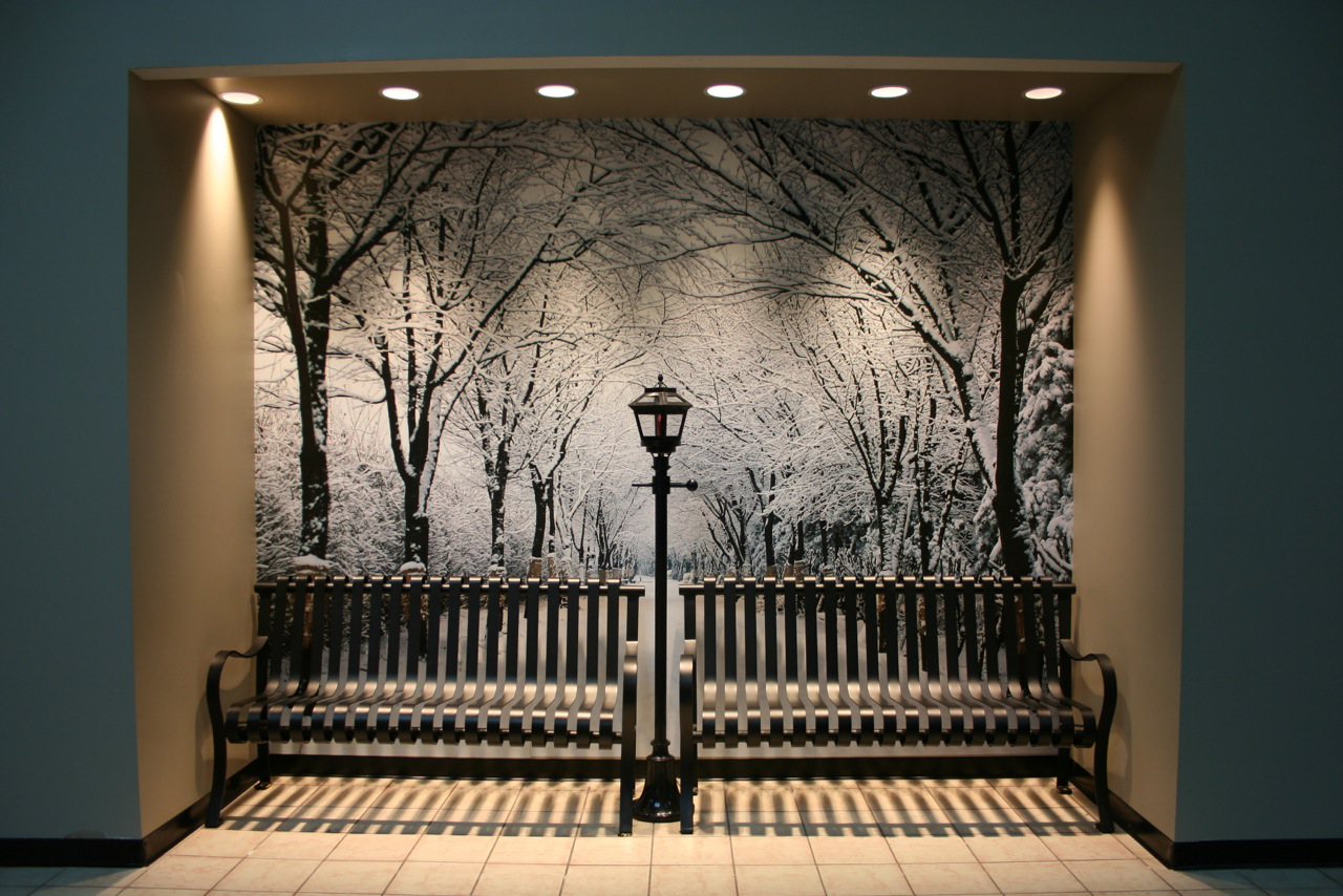Two black metal benches separated by a lamp post in front of a wall mural showing a snow-covered tree-lined pathway.