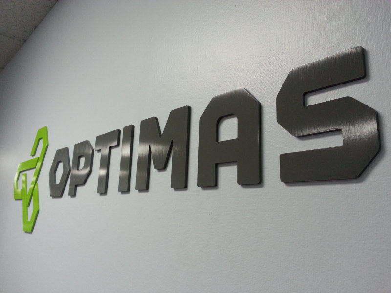 Wall-mounted 3D sign with a green geometric logo and the word OPTIMAS in large black letters.
