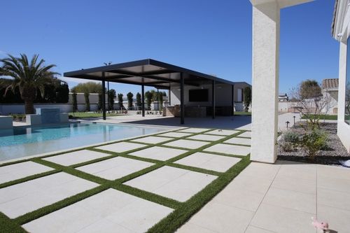 Modern backyard with tiled patio, grassy grid pattern, swimming pool, palm tree, and covered outdoor lounge.