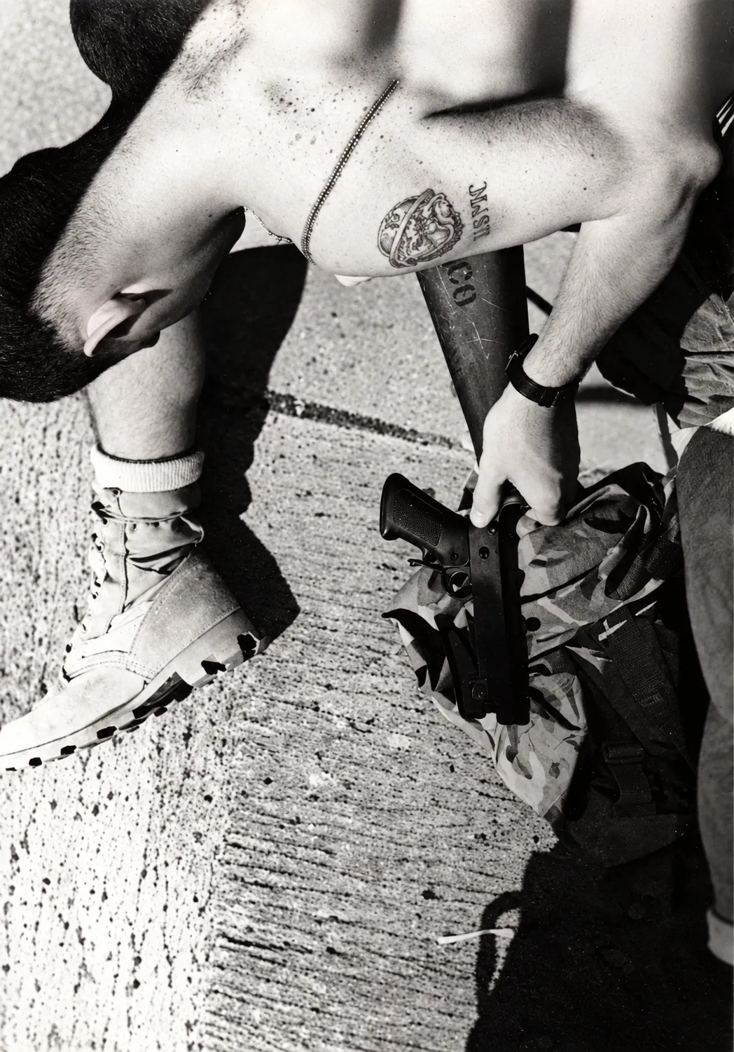 Black-and-white photograph of a seated person with a tattoo and military equipment.