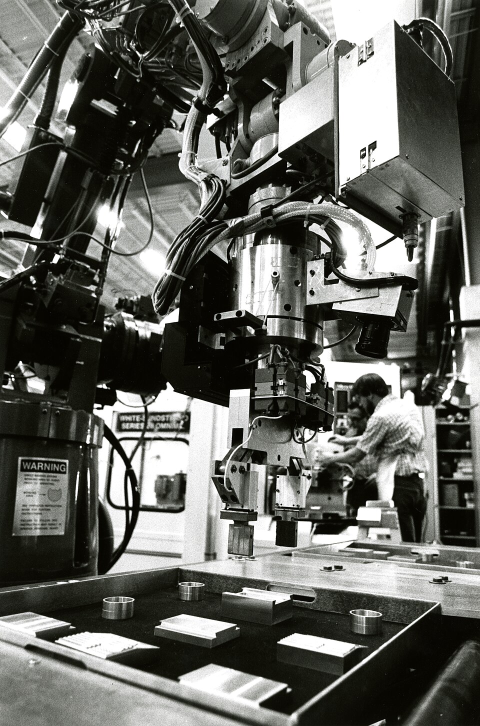 Industrial robotic gripper above machined parts, with a technician working in the background.
