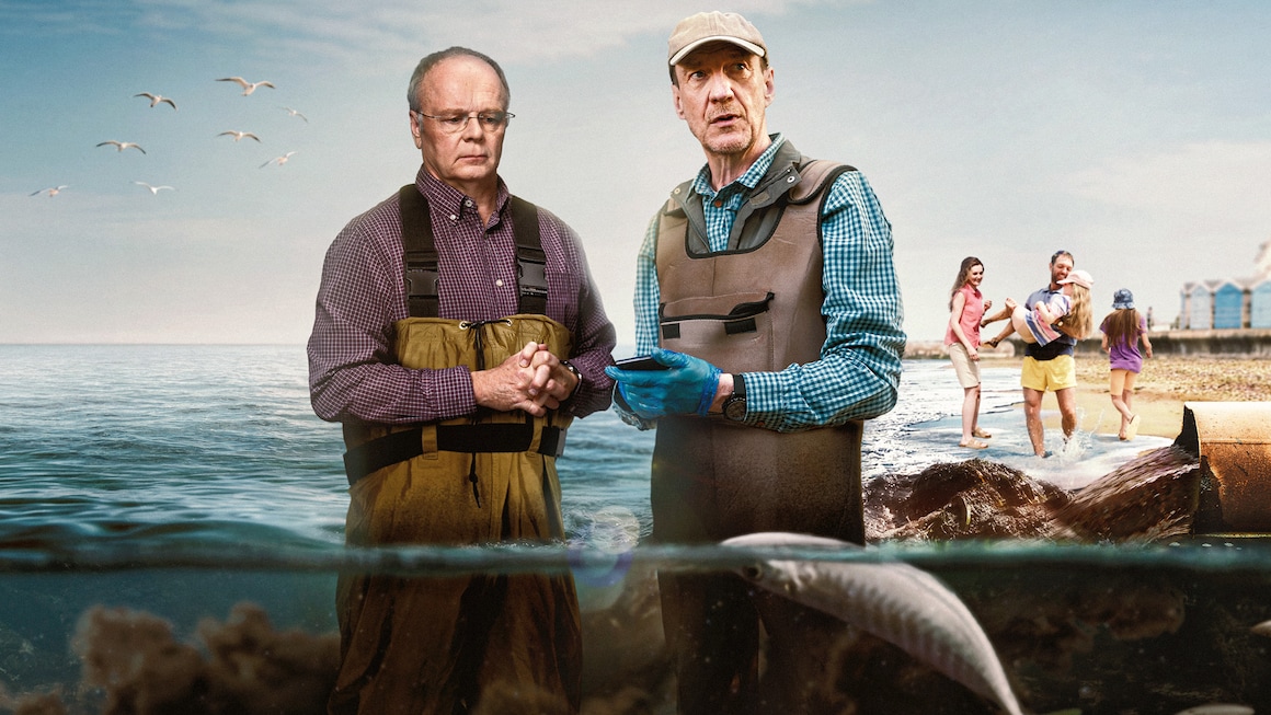 Two men stand in the sea surrounded by sewage, with a family playing on the shore behind them.