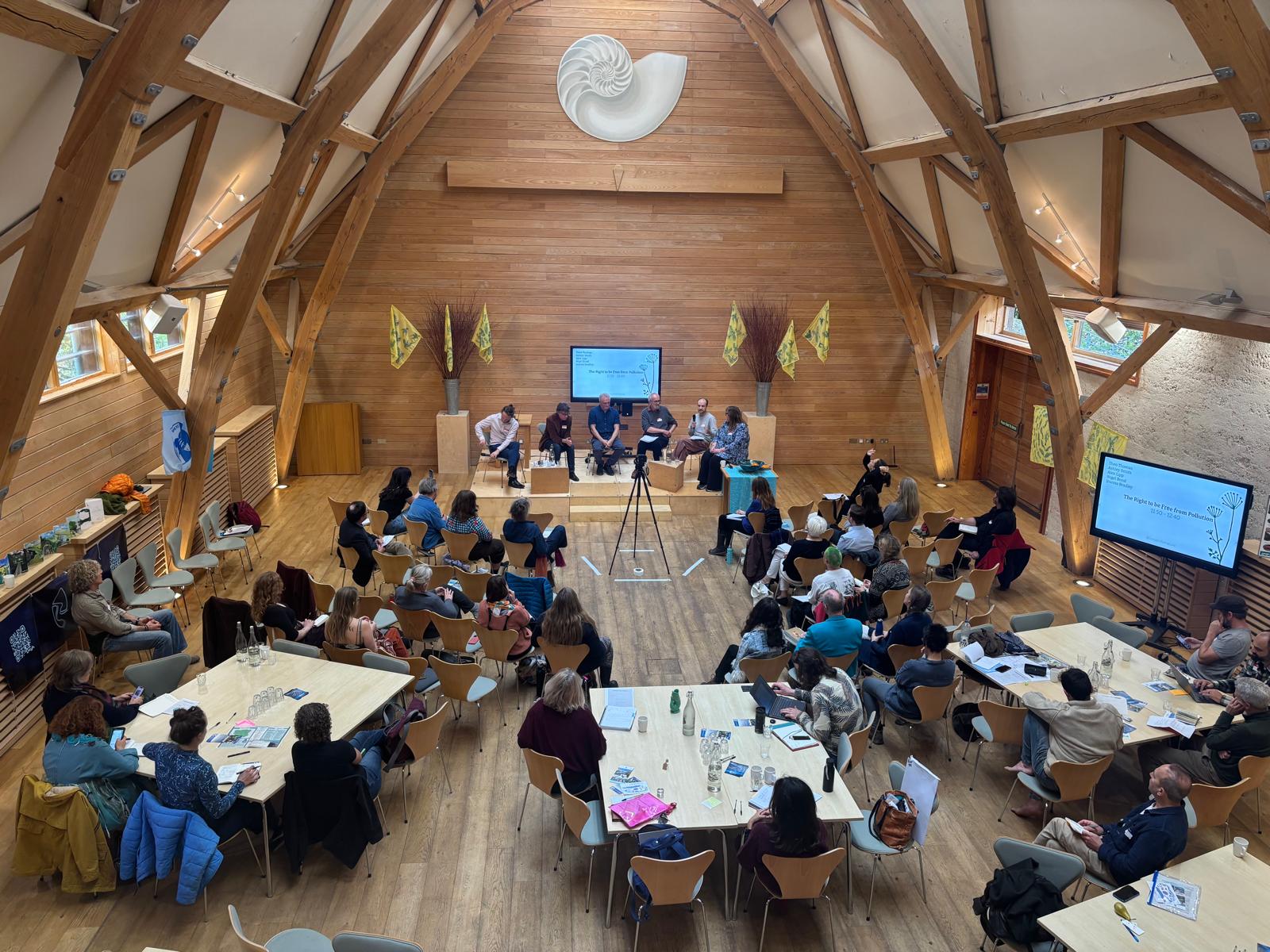People sat on chairs around tables in front of a stage on which is sat a panel of six people. 