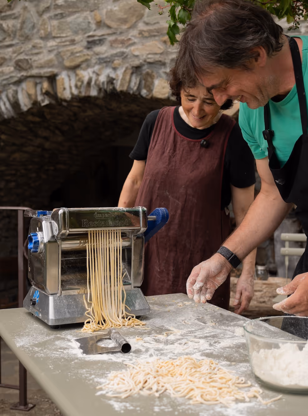 Zwei Menschen, die mit einer Nudelmaschine frische Pasta herstellen, umgeben von Mehl auf einem Tisch.