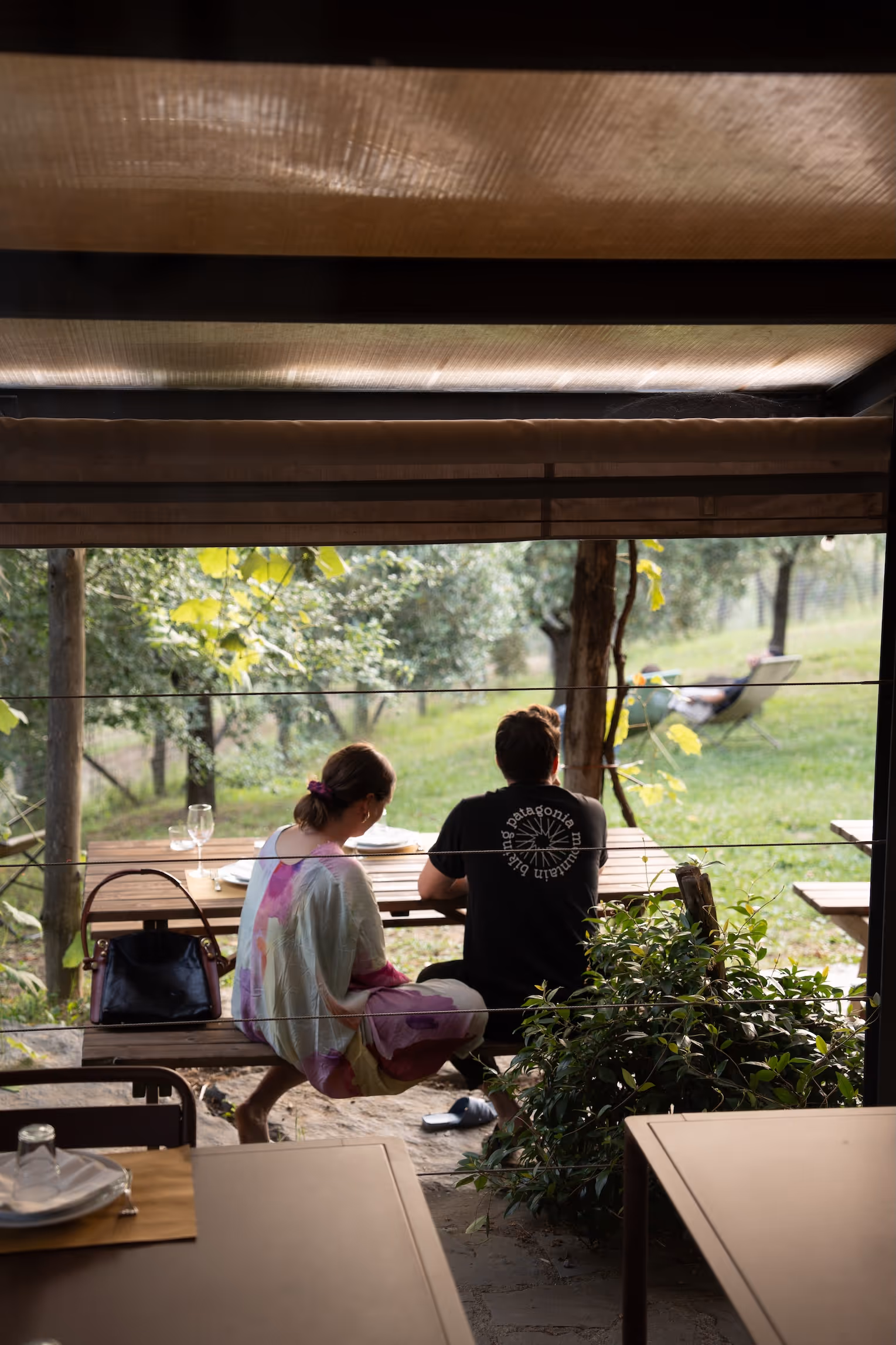 Zwei Personen sitzen auf einer Holzbank an einem Tisch unter einem Überdach im Garten mit grüner Landschaft im Hintergrund.