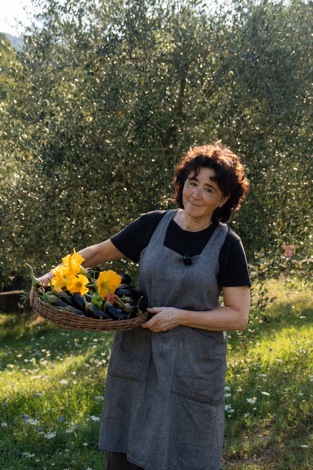 Frau in grauer Schürze hält einen Korb mit frischem Gemüse und gelben Blüten in einem sonnigen Garten.
