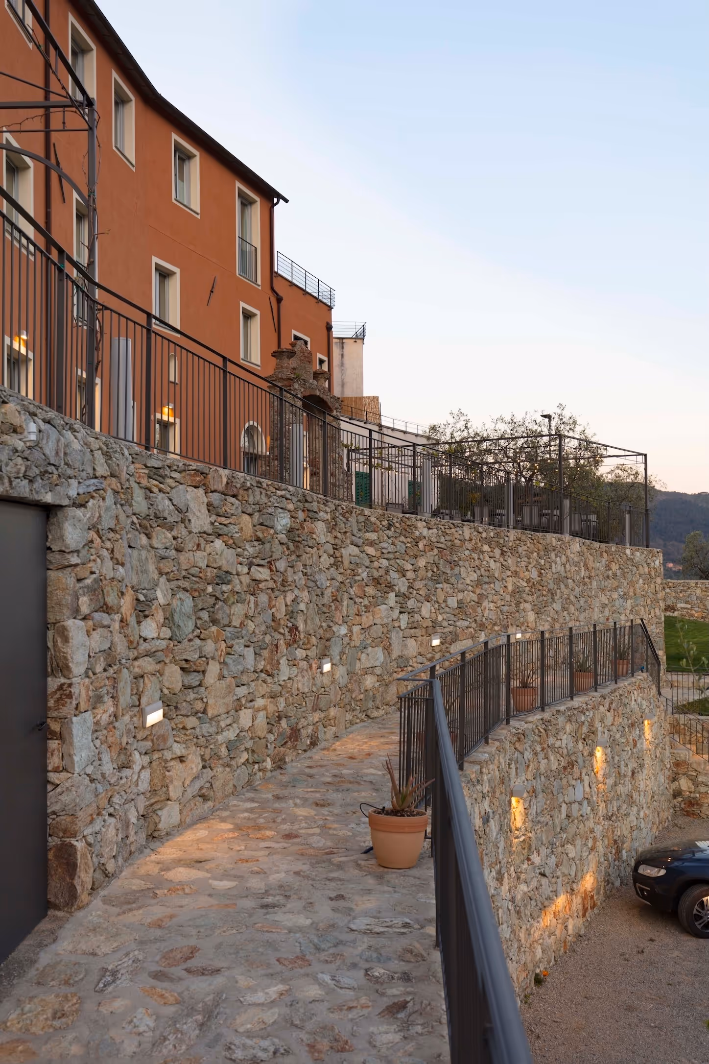 Gepflasterter Weg entlang einer Steinmauer mit Metallgeländer neben einem orangen Gebäude unter hellem Himmel.