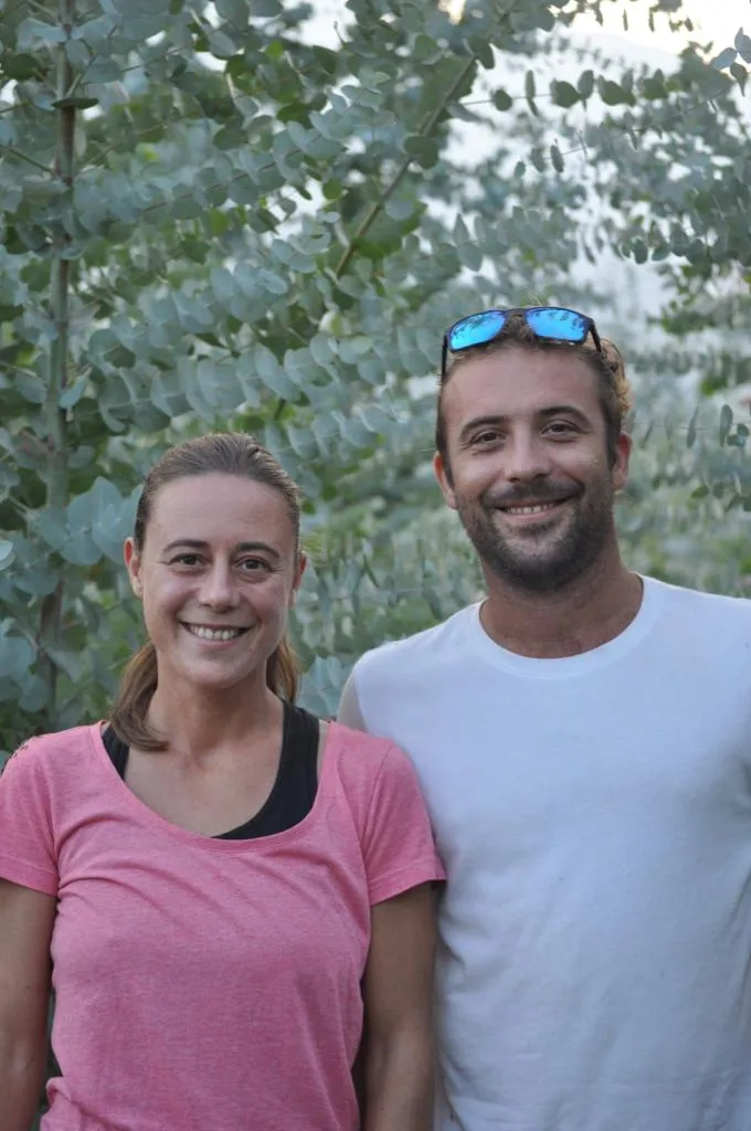 Ein lächelndes Pärchen steht vor einem grünen Pflanzenhintergrund; die Frau trägt ein rosa T-Shirt, der Mann ein weißes T-Shirt mit Sonnenbrille auf dem Kopf.