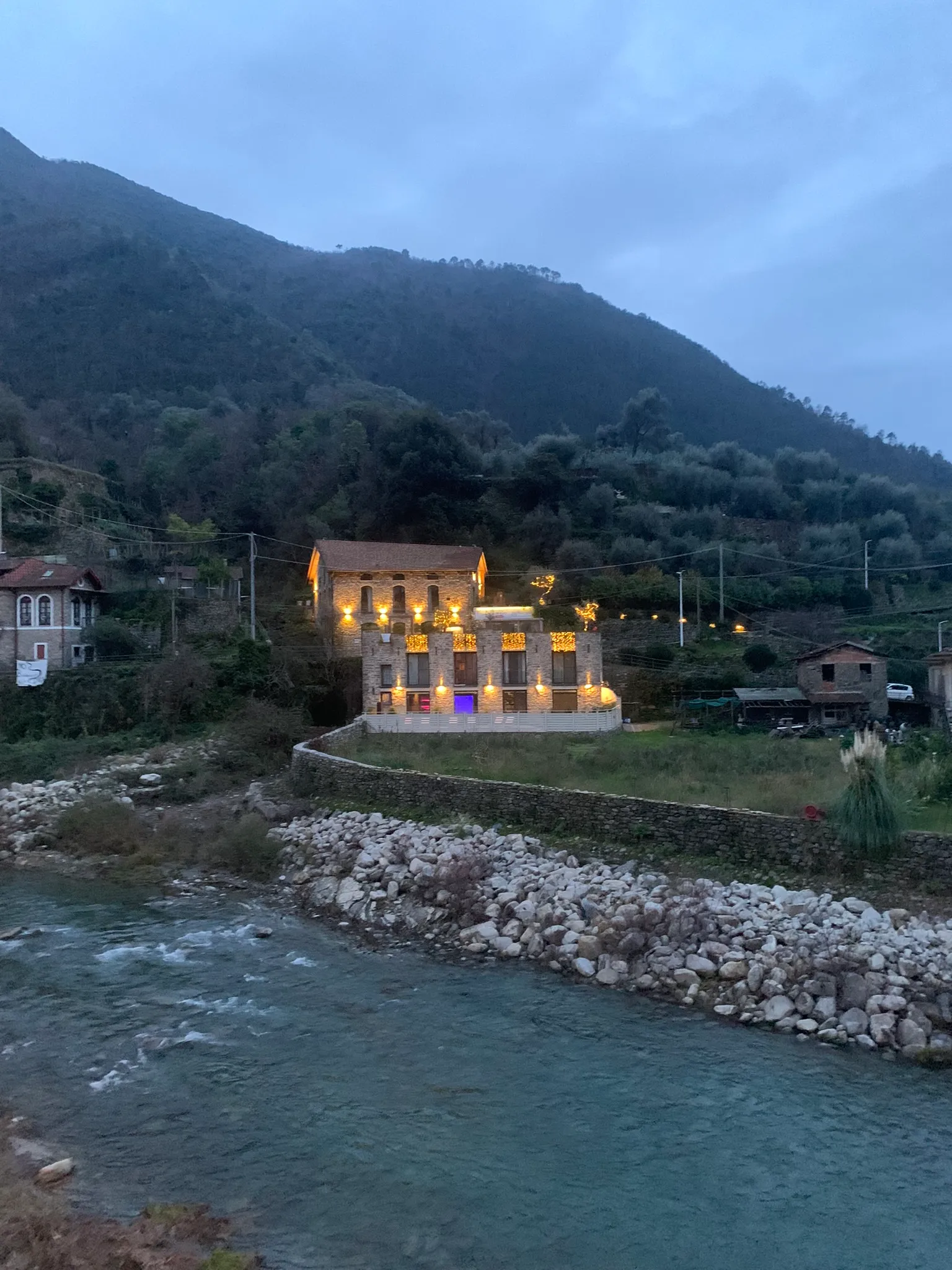 Beleuchtetes Steinhaus am Flussufer vor bewaldetem Hügel bei Dämmerung.