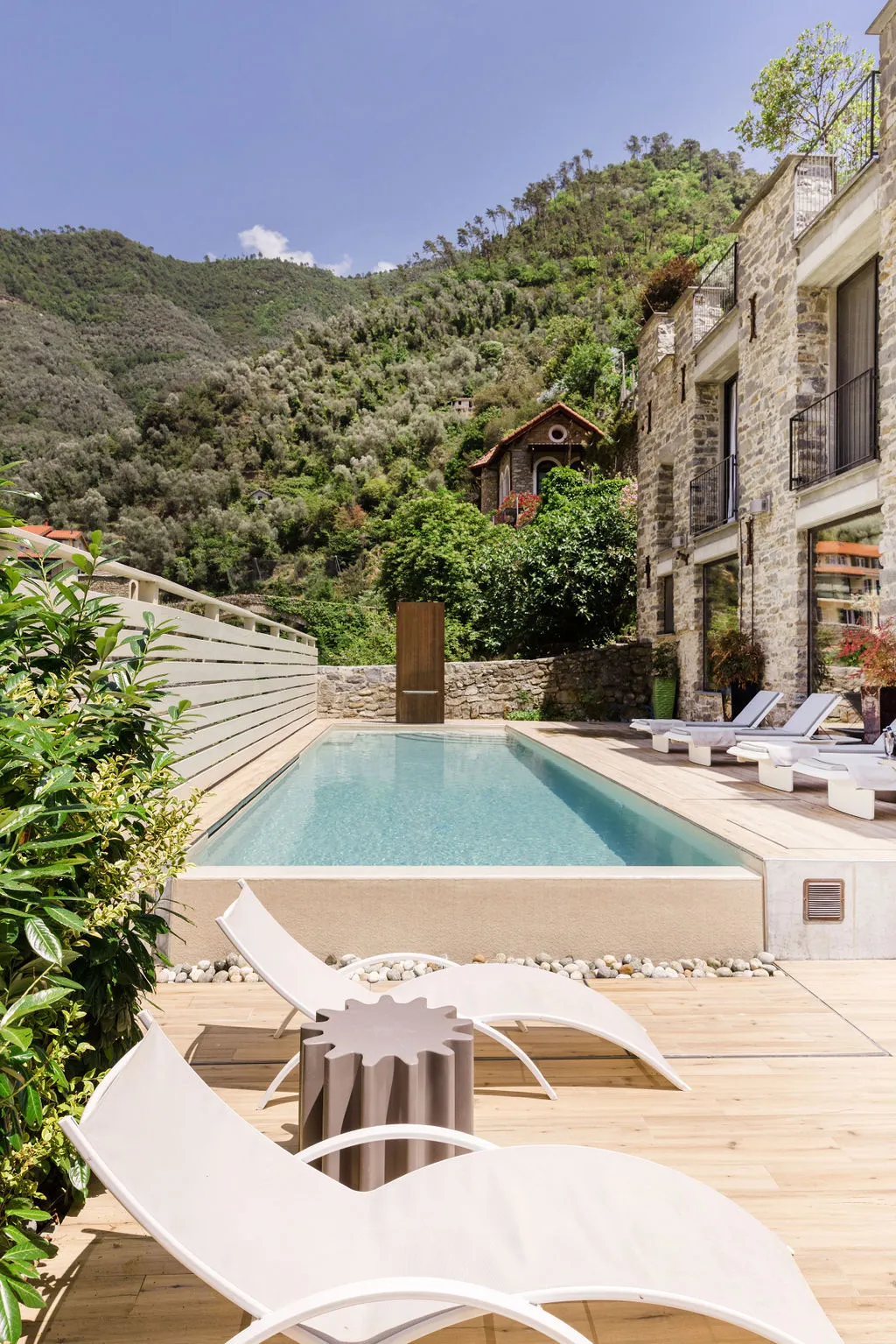 Moderner Pool mit Liegestühlen auf Holzdeck vor einem Natursteinhaus, umgeben von grünen Hügeln.