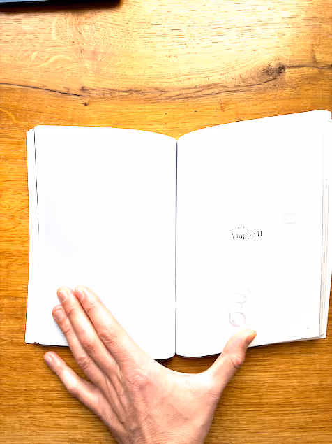 Eine Hand hält ein aufgeschlagenes Buch mit weißen Seiten über einem Holztisch.