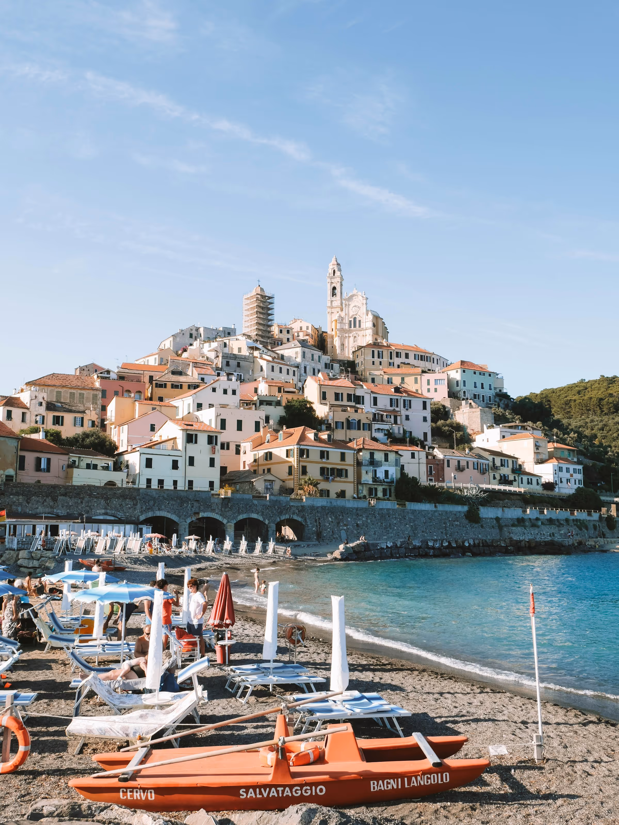 Italienisches Küstendorf mit bunten Häusern am Hang, Strand mit Sonnenliegen und einem roten Rettungsboot.