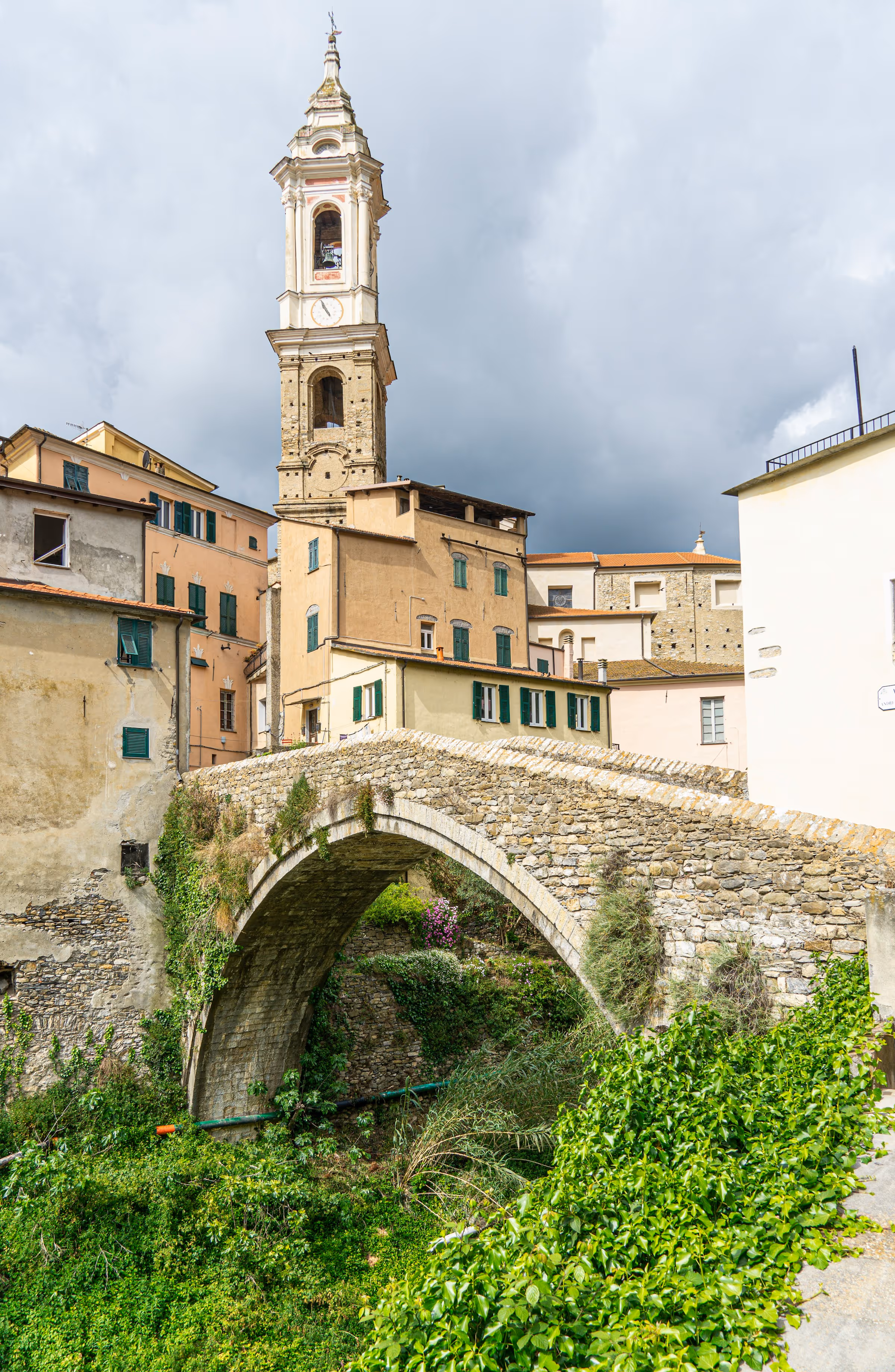Steinerne Bogenbrücke mit Bewuchs und dahinter historische Gebäude mit Kirchturm unter bewölktem Himmel.