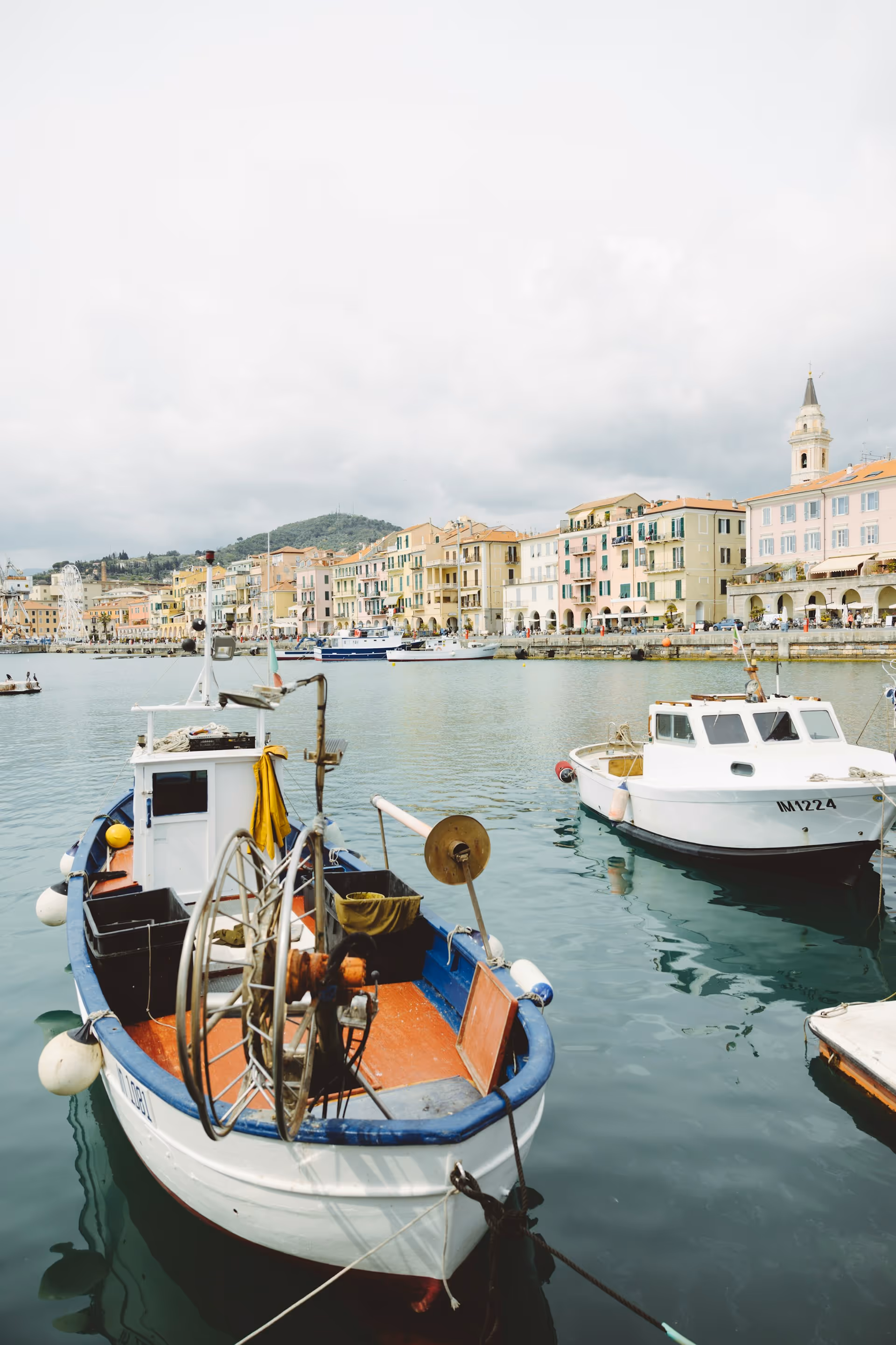 Boote im Hafen von Imperia Oneglia mit pastellfarbenen Gebäuden und einer Kirchturmspitze im Hintergrund.