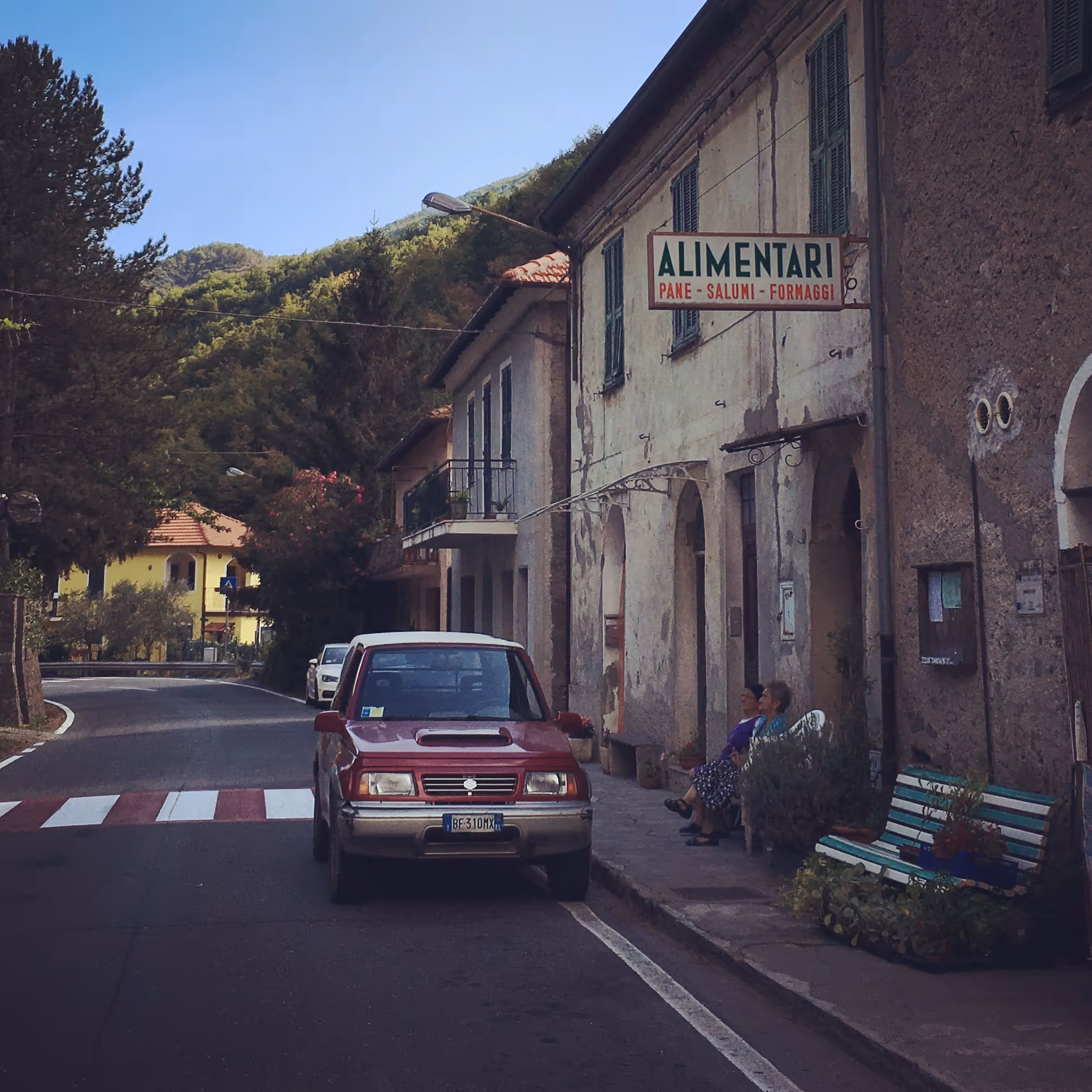 Altes italienisches Lebensmittelgeschäft an einer Straße mit rot-weißem Zebrastreifen und einem geparkten roten Auto, zwei ältere Frauen sitzen auf einer Bank davor.