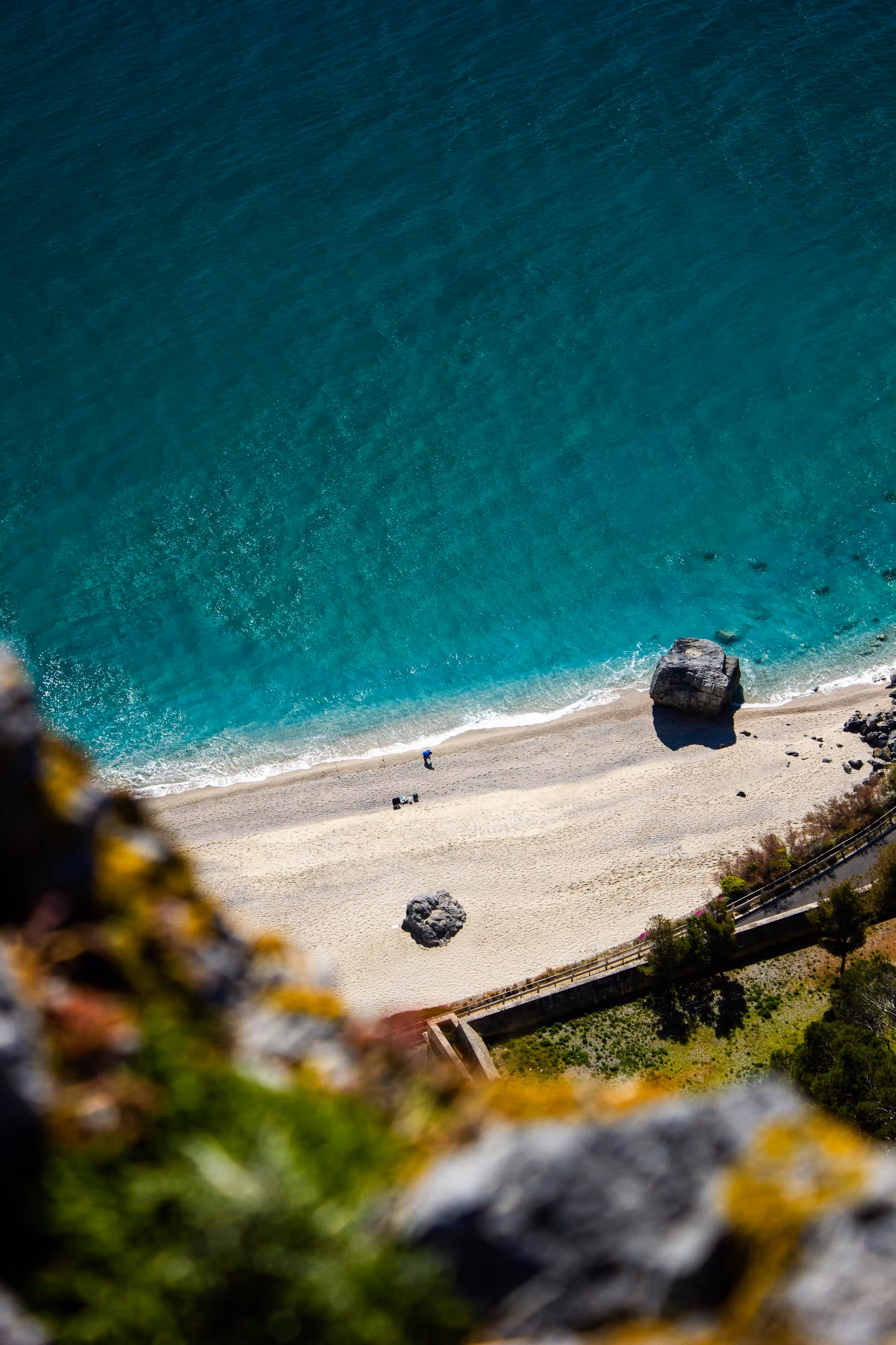 Luftaufnahme eines ruhigen Strandes mit türkisfarbenem Wasser, zwei großen Felsen im Sand und einer Person, die am Ufer spazieren geht.