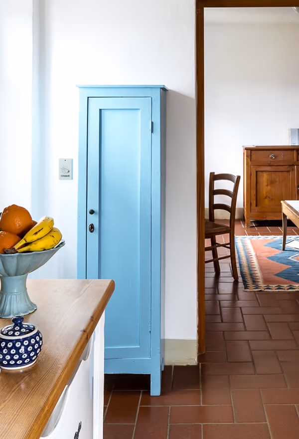 Ein heller Raum mit einem kleinen, blauen Schrank an der Wand, links davon ein Holztisch mit einer Schale voller Orangen und Bananen sowie einer kleinen blauen Keramikschale. Der Fliesenboden ist terrakottafarben, im Hintergrund ein weiterer Raum mit einem Holzstuhl, einem Teppich mit geometrischem Muster und einem Holzmöbel.