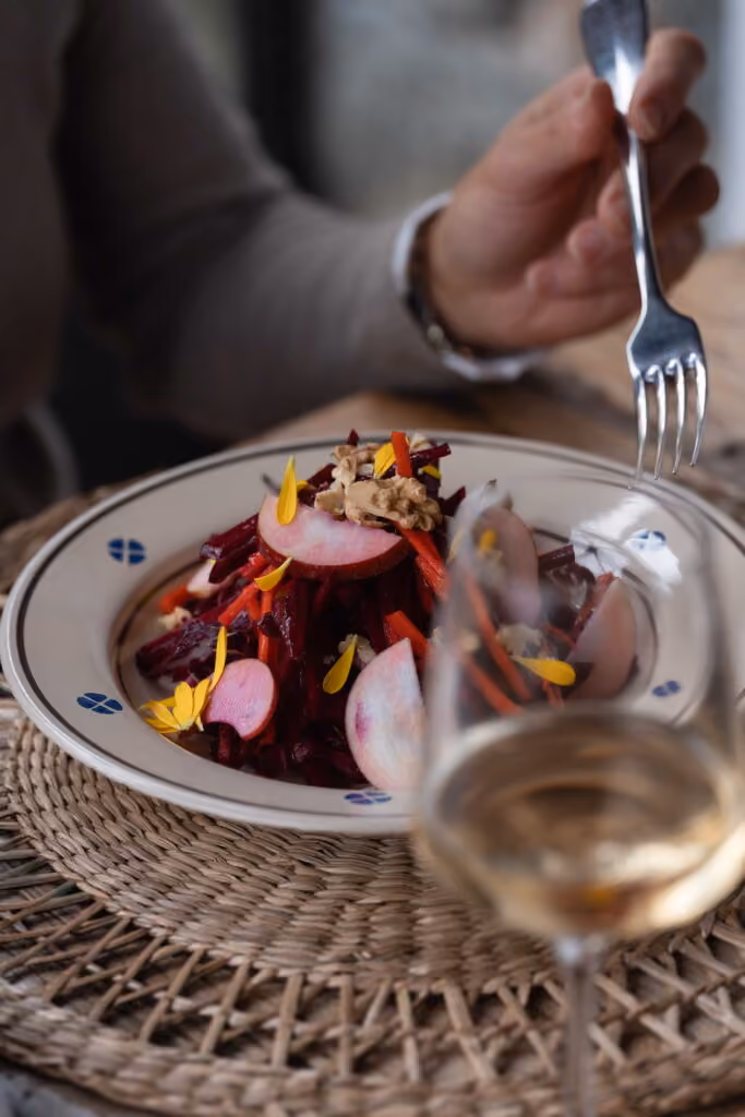 Nahaufnahme eines bunt garnierten Salats mit roten Rüben, Radieschen, Karottenstreifen, Walnüssen und essbaren Blüten auf einem weißen Teller, daneben ein Glas Weißwein und eine Person mit Gabel.