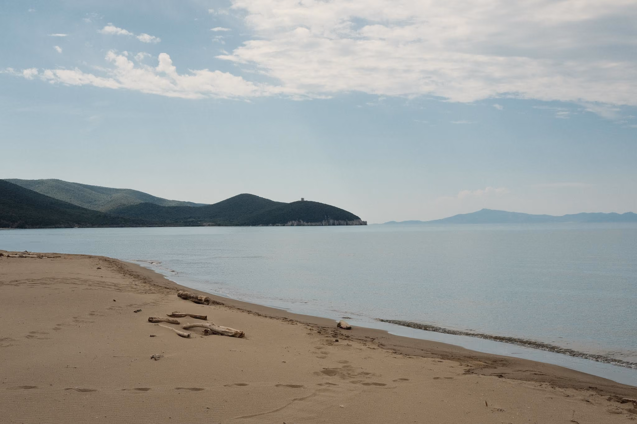 Ein ruhiger Sandstrand mit Fußspuren und wenigen Treibholzstücken am Ufer eines stillen Meeres, im Hintergrund bewaldete Hügel und ein teilweise bewölkter Himmel.