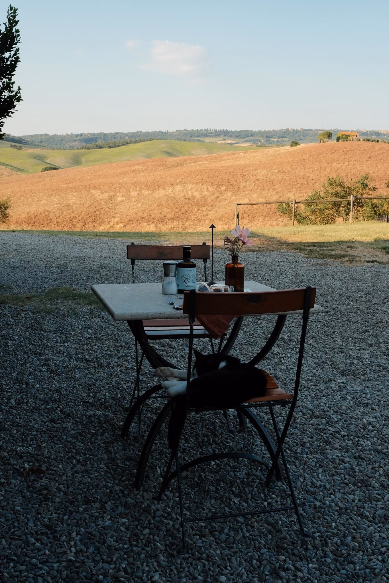 Kleiner Außentisch mit zwei Stühlen auf Kies vor einer weiten Landschaft mit grünen und braunen Hügeln unter blauem Himmel.