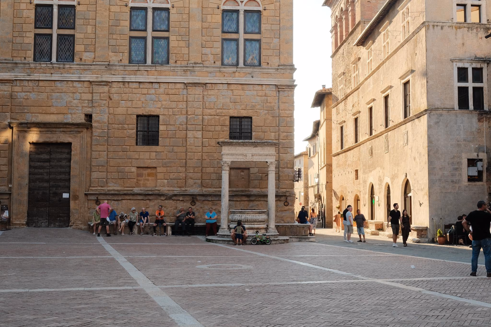 Platz mit historischem Brunnen und Steinmauer mit großen Fenstern, umgeben von alten Gebäuden, mit Leuten, die sich auf einer Bank ausruhen und spazieren gehen.
