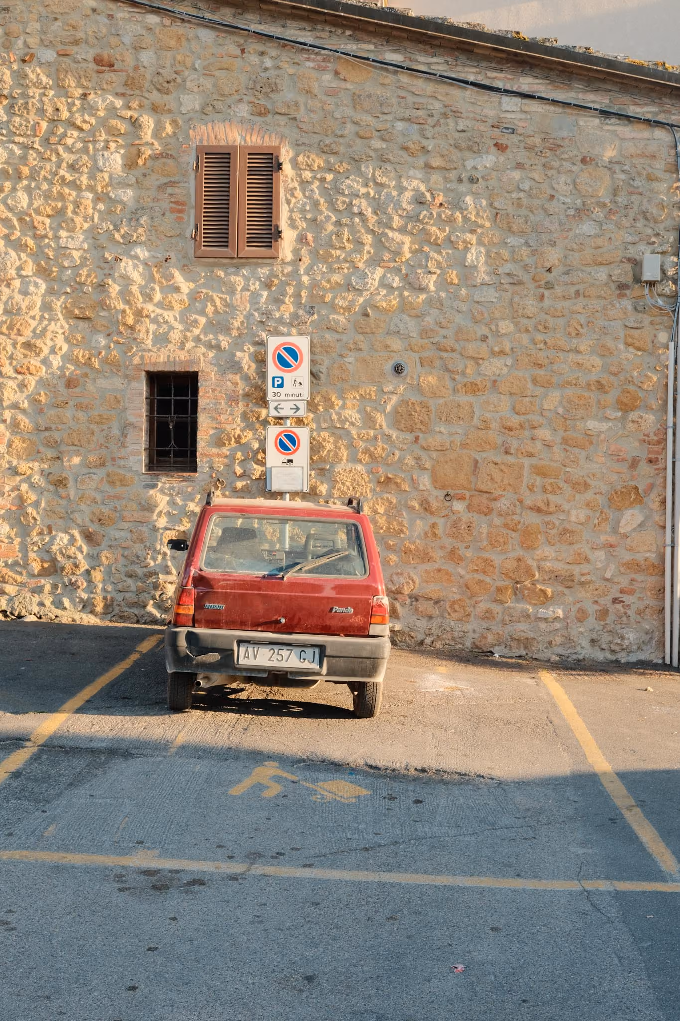 Roter alter Fiat Panda geparkt in einer Parklücke vor einer Steinmauer mit zwei Fenstern und Verkehrsschildern, die Parkbeschränkungen anzeigen.