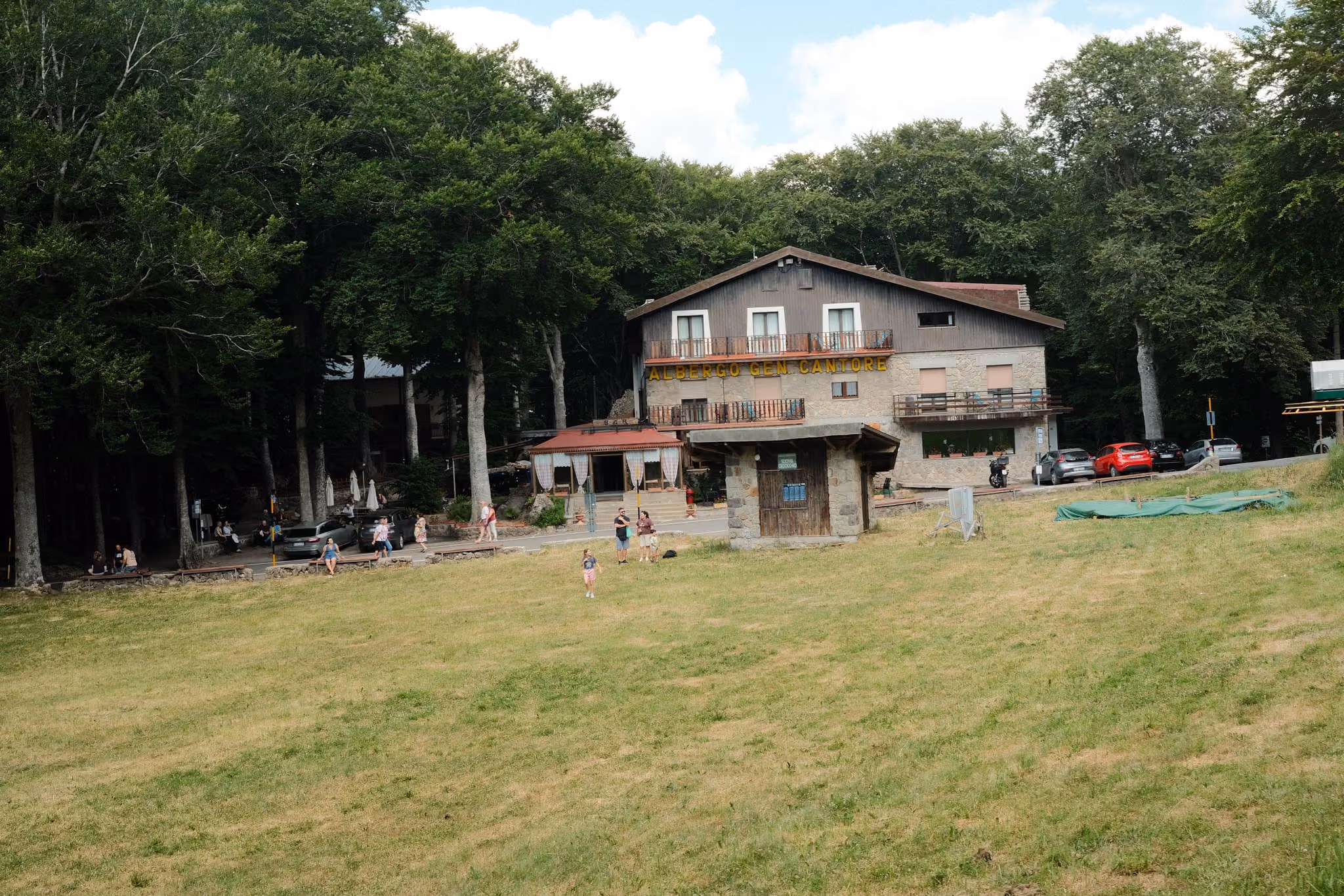 Ein großes, zweistöckiges Holz- und Steinhaus namens Albergo Gen Cantore umgeben von Bäumen in einer grünen Wiese mit mehreren Menschen, die sich im Freien aufhalten.