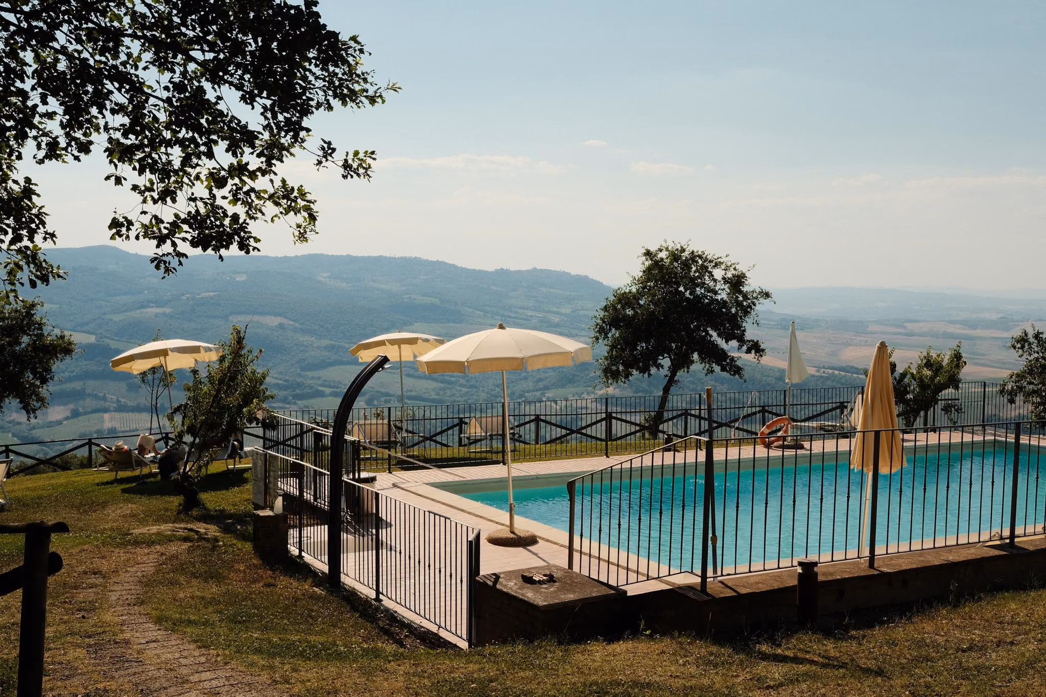 Ein Pool mit blauem Wasser und Sonnenschirmen auf einem Hügel, umgeben von einem schwarzen Zaun, mit Blick auf sanfte grüne Hügel und einen bewölkten Himmel im Hintergrund.